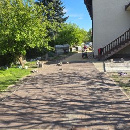 BRUKART - Brązowa kostka brukowa ułożona częściowo na podjeździe. W tle dom, drzewa i pracownik z zagęszczarką. Na pierwszym planie cień drzewa na kostce.