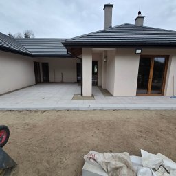 BRUKART - Nowoczesny taras z jasnych, kwadratowych płyt, częściowo ułożony przed domem. Widoczny wózek budowlany i worki z materiałami. Pochmurne niebo w tle.