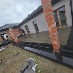 BRUKART - Montaż tarasu z kamiennych płyt przy domu jednorodzinnym. Widoczny mur z cegieł, częściowo ułożone płyty i pracownik z miarką. Realizacja w pochmurny dzień.