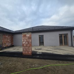 BRUKART - Budowa tarasu z czarnych płyt, widoczne ceglane słupy i przygotowane podłoże z piasku. W tle fragment domu w stanie surowym zamkniętym, pochmurne niebo.