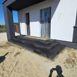 BRUKART - Nowoczesne schody kamienne z ciemnego granitu w Gorzkowicach, prowadzące do wejścia budynku. Widoczne świeżo ułożone stopnie i piasek. Budynek w bieli i szarości.