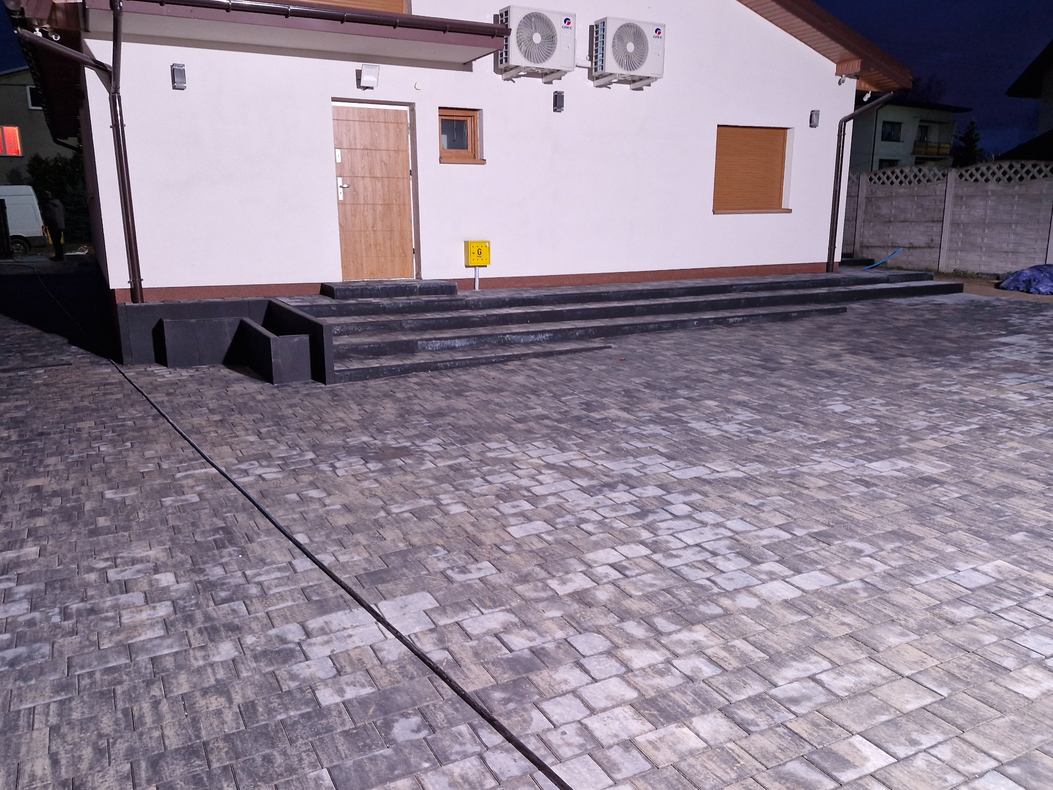Szara kostka brukowa ułożona przed domem z szarymi schodami wejściowymi i dwoma klimatyzatorami na ścianie, widok wieczorny.