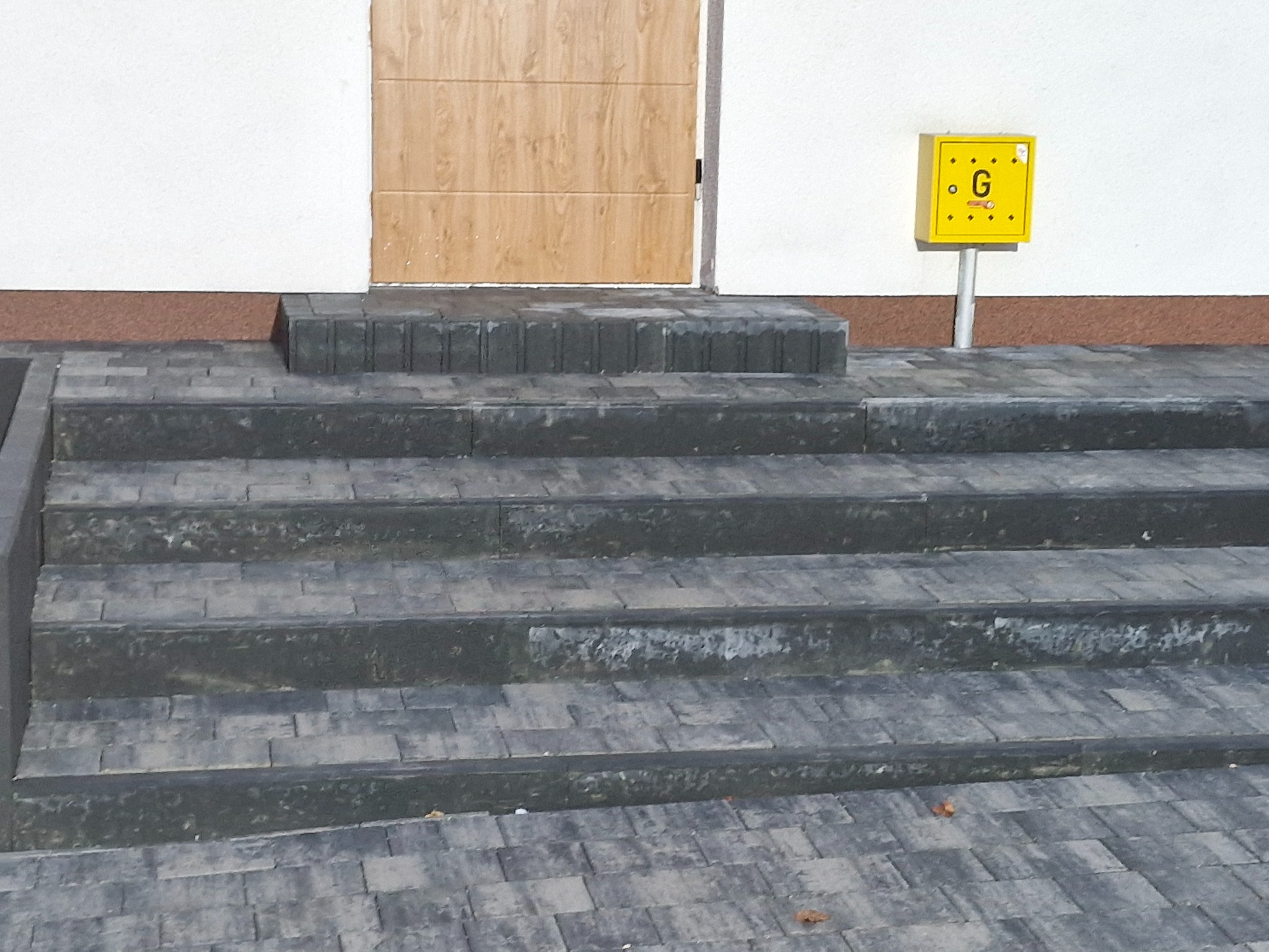 Schody wejściowe z szarej kostki brukowej prowadzące do drzwi garażowych, obok żółta skrzynka gazowa.