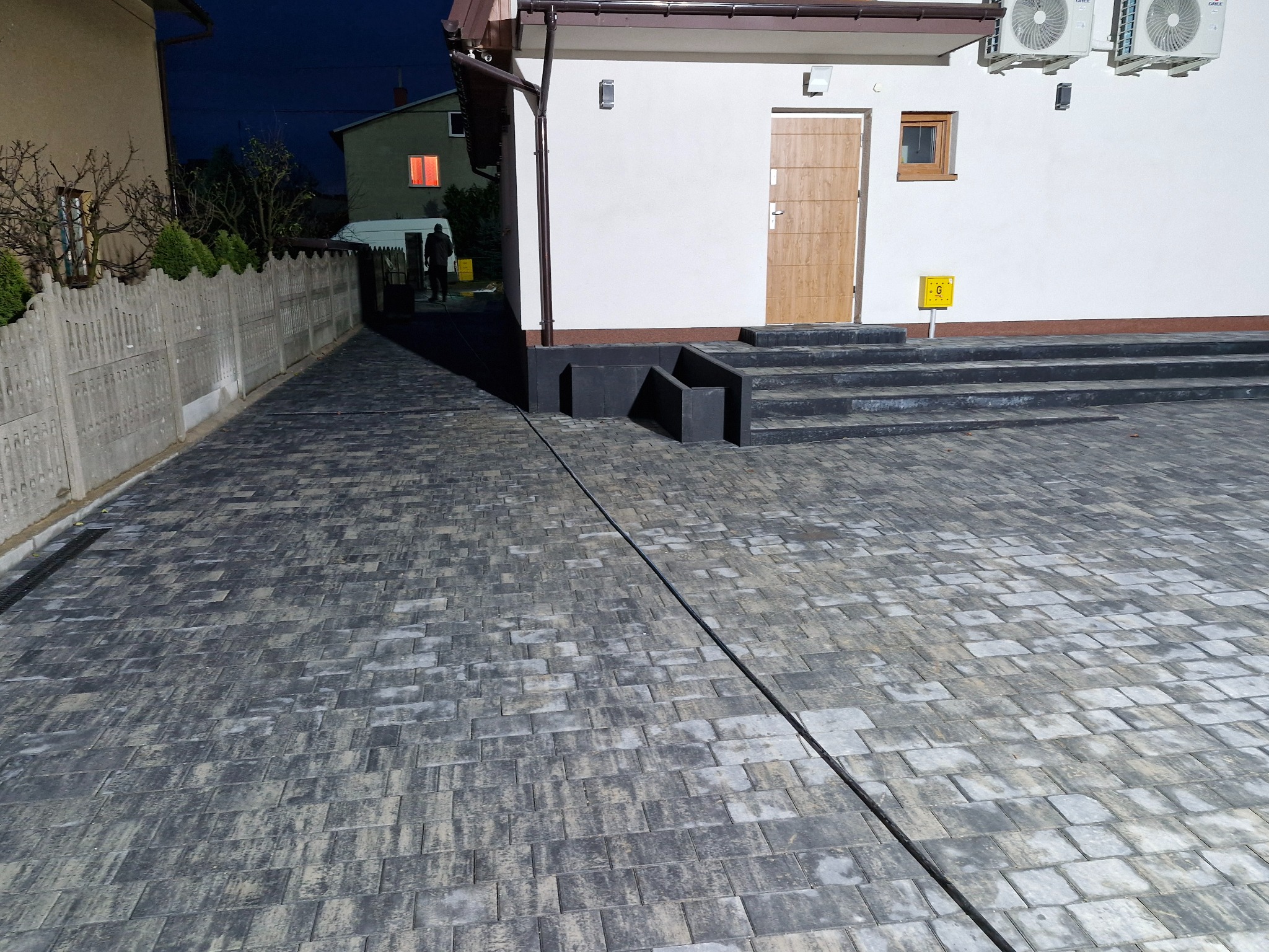 Ułożona szara kostka brukowa z odpływem liniowym przed budynkiem z kamiennymi schodami wejściowymi i dwoma klimatyzatorami na ścianie, widoczne światło w oknie sąsiedniego domu w nocy.