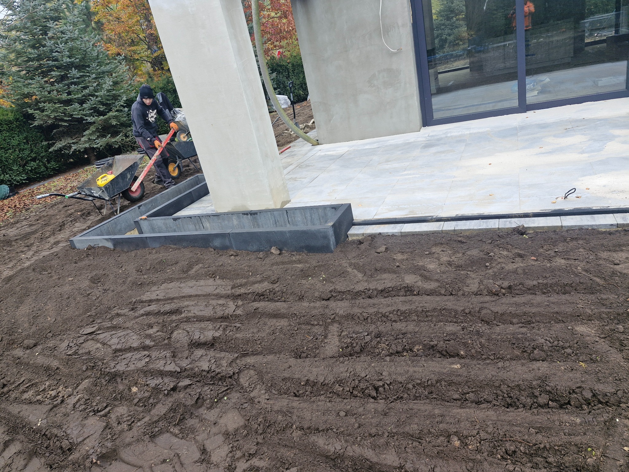 Pracownik z taczką i poziomicą przygotowuje obrzeże tarasu z szarych betonowych krawężników na ubitej ziemi przed nowoczesnym domem z dużymi oknami.