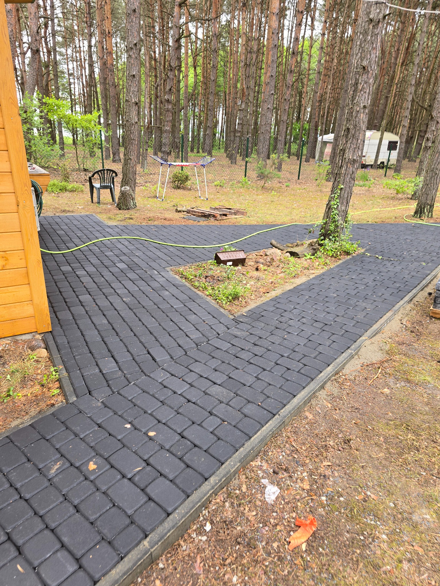 Ścieżka z ciemnej kostki brukowej, wijąca się między drzewami na posesji. Widoczne obrzeża, trawa i leśne otoczenie z domkiem letniskowym w tle.