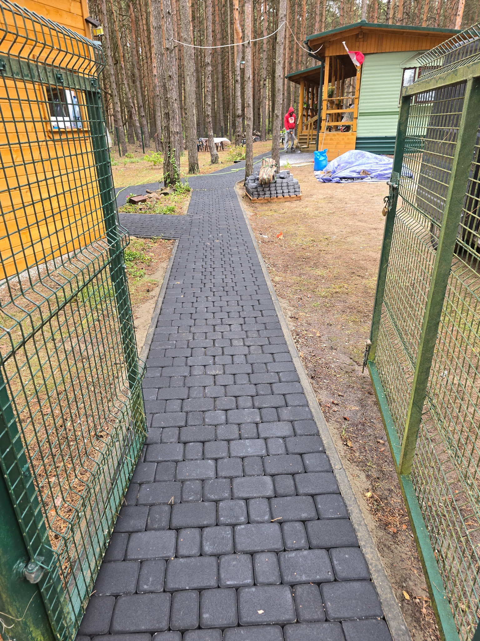 Ścieżka z ciemnej kostki brukowej prowadzi przez otwartą, zieloną furtkę w kierunku domku w lesie. Ułożona kostka tworzy równy chodnik.