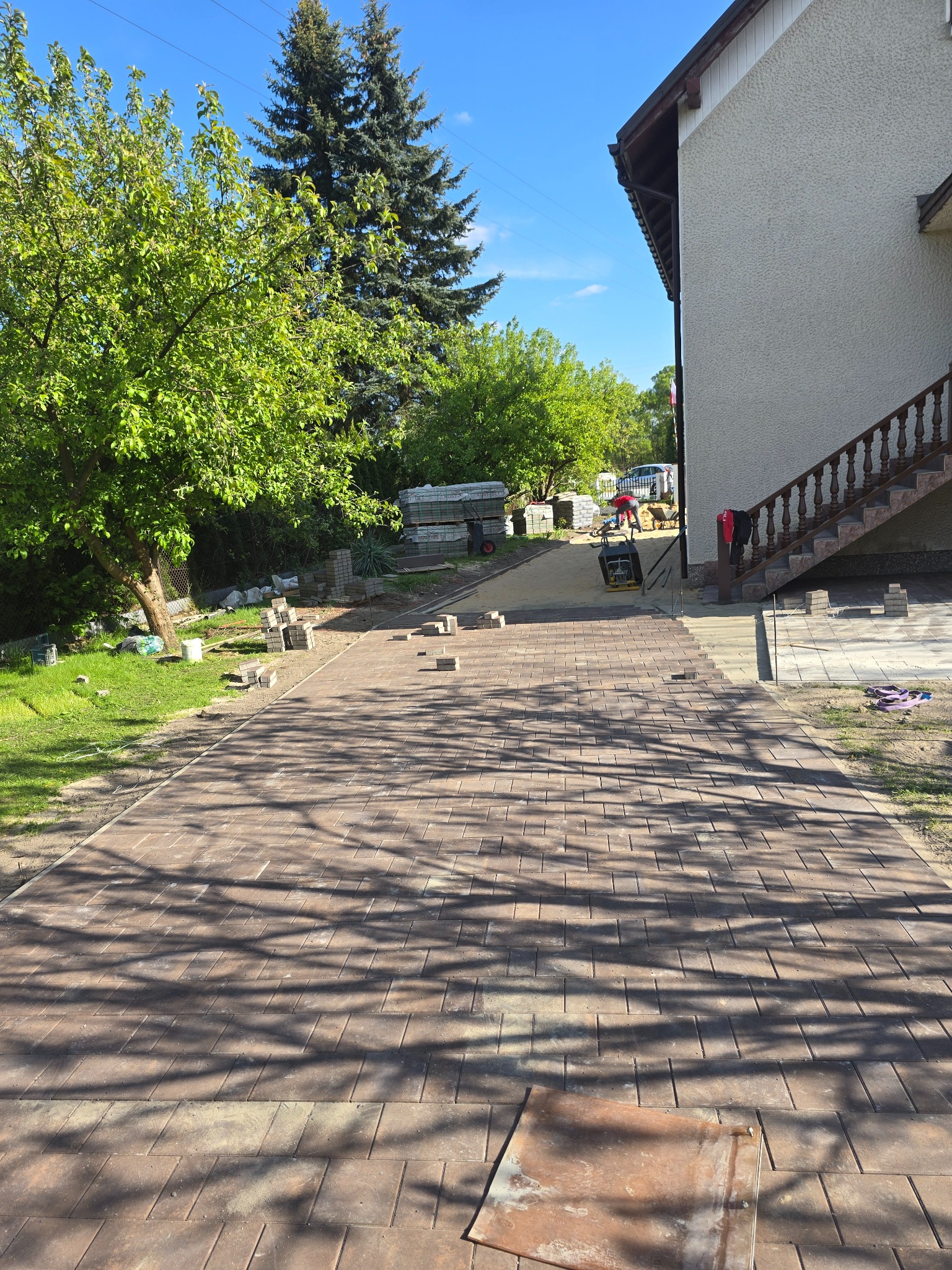 Brązowa kostka brukowa ułożona częściowo na podjeździe. W tle dom, drzewa i pracownik z zagęszczarką. Na pierwszym planie cień drzewa na kostce.