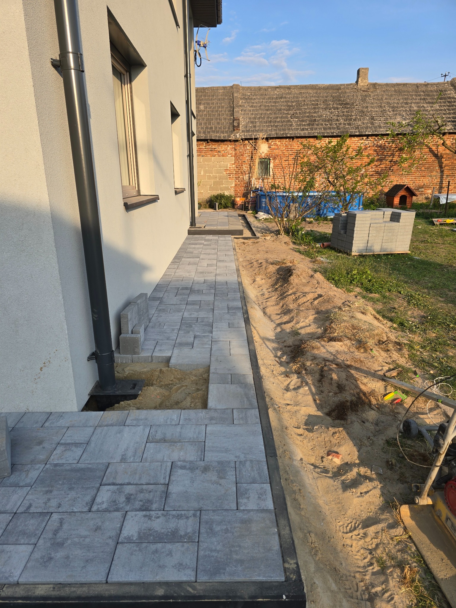 Fragment układanej ścieżki z szarych kostek brukowych przy budynku. Część kostek jeszcze nieułożona, widoczna ziemia i paleta z kostką w tle.