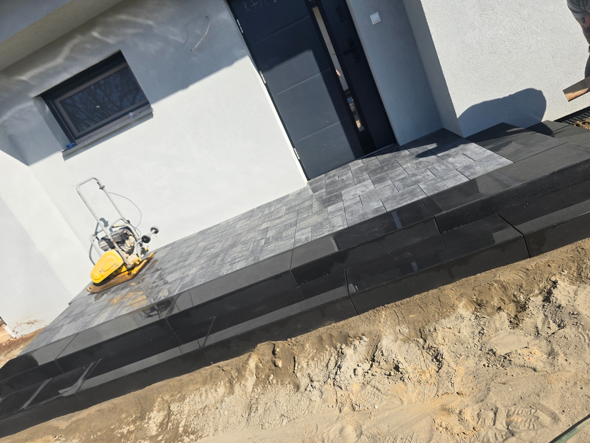 Nowoczesne schody wejściowe z kostki brukowej w odcieniach szarości i czerni, częściowo ułożone, z zagęszczarką w tle, w trakcie prac wykończeniowych.