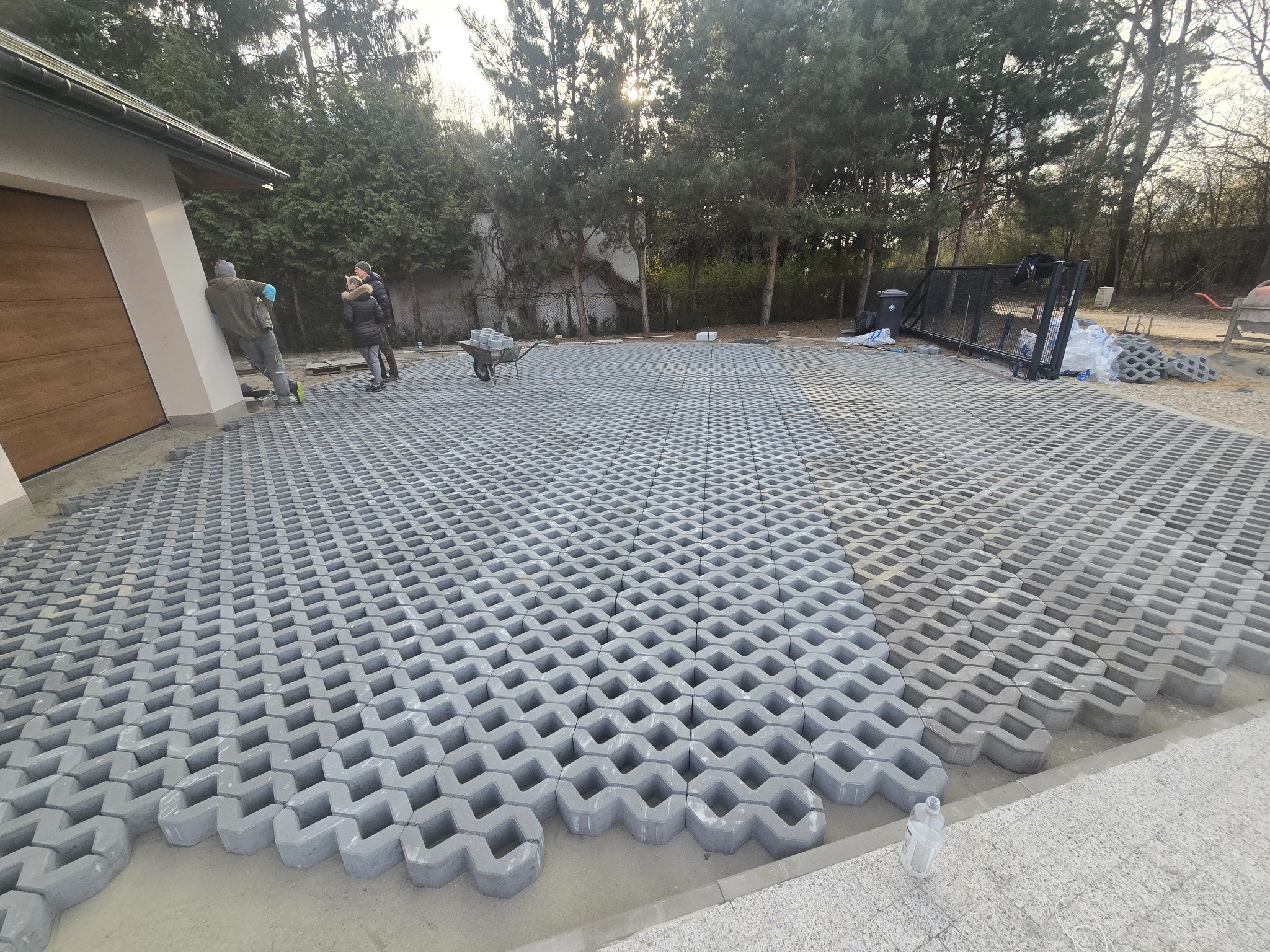 Ułożona szara ażurowa kostka brukowa na podjeździe, z widocznymi pracownikami i taczką z kostką w tle. Oświetlenie słoneczne, drzewa w tle.