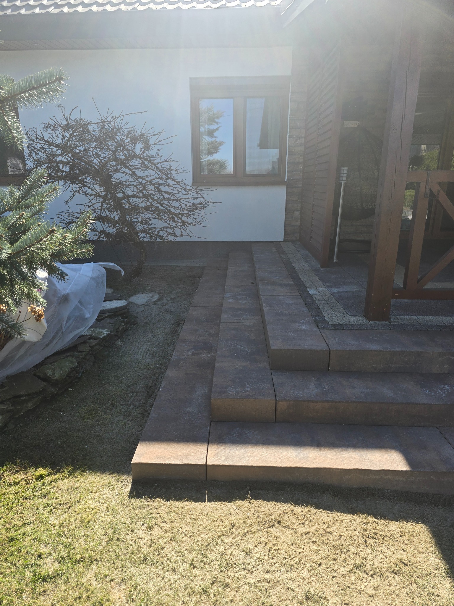 Schody z kostki brukowej prowadzące do wejścia domu, z elementami trawnika i roślinności w tle. Widoczne detale wykończenia i oświetlenie słoneczne.