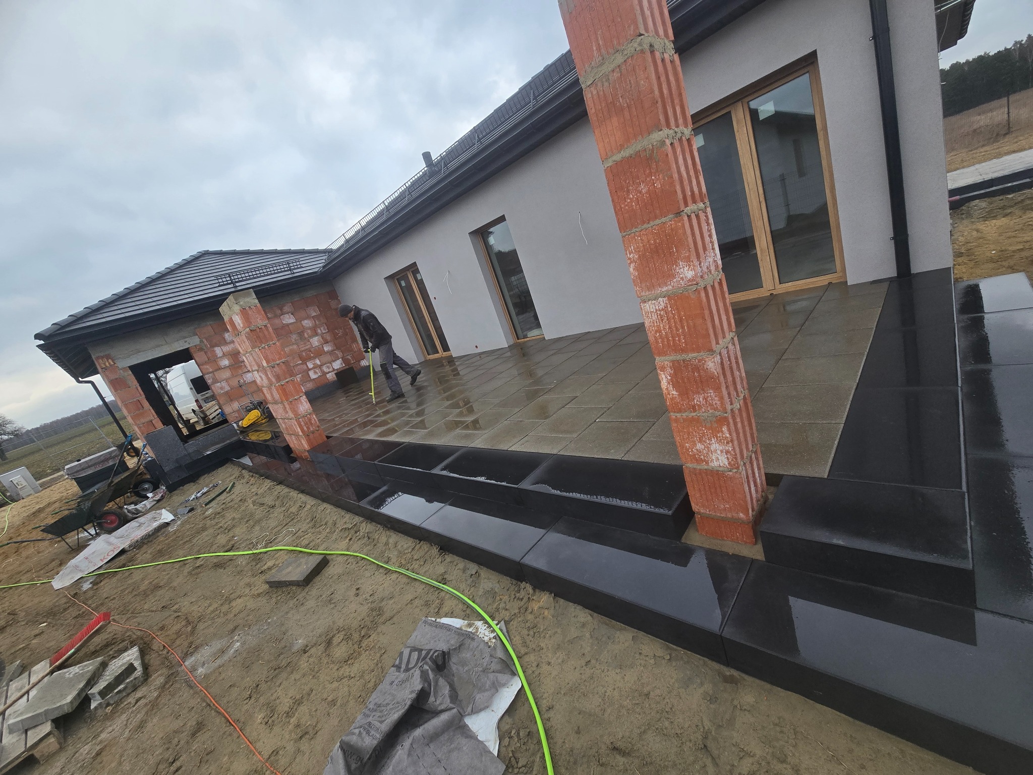 Montaż tarasu z kamiennych płyt przy domu jednorodzinnym. Widoczny mur z cegieł, częściowo ułożone płyty i pracownik z miarką. Realizacja w pochmurny dzień.