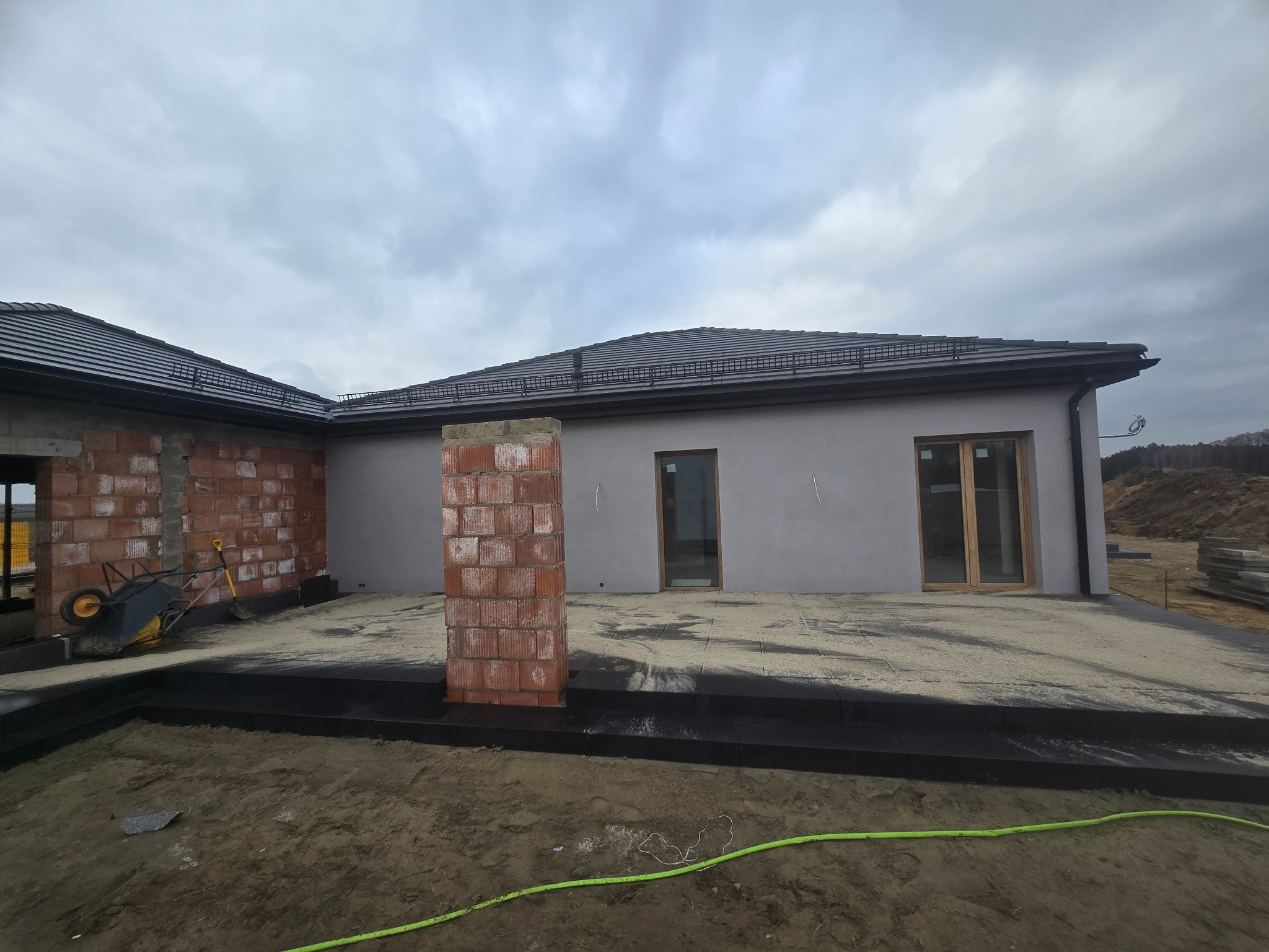 Budowa tarasu z czarnych płyt, widoczne ceglane słupy i przygotowane podłoże z piasku. W tle fragment domu w stanie surowym zamkniętym, pochmurne niebo.