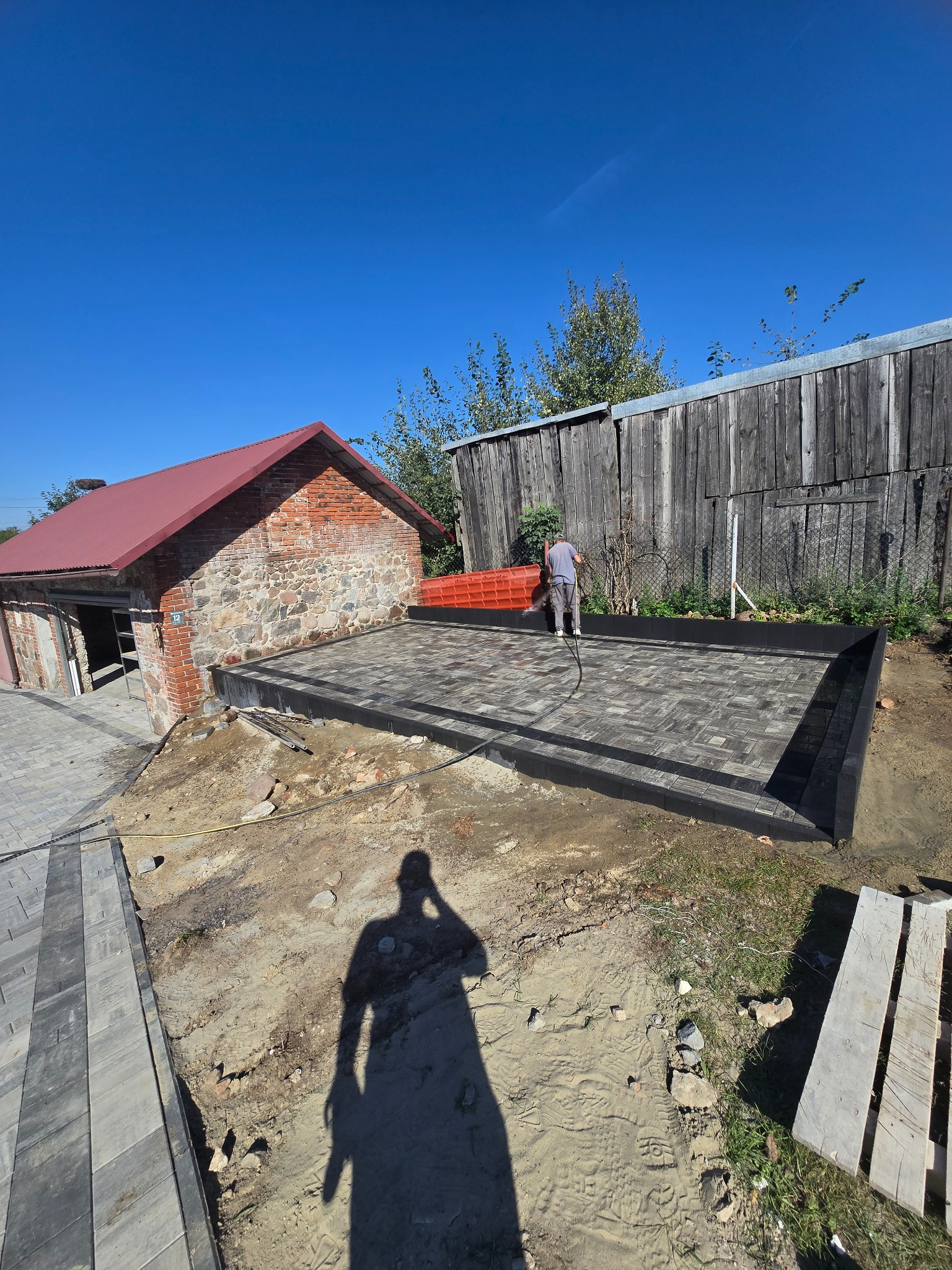 Układanie kostki brukowej na posesji. Widoczny fragment ułożonej nawierzchni, pracownik z wężem i budynek gospodarczy z czerwonej cegły w tle.