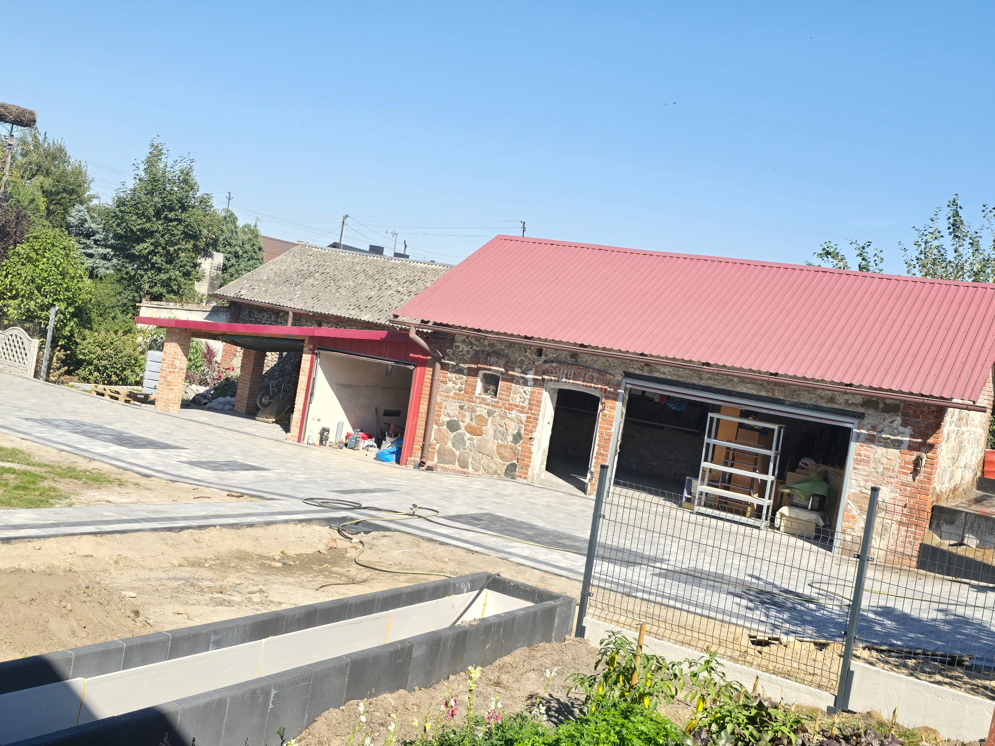 Nowa, jasnoszara kostka brukowa ułożona na podjeździe i chodniku, obok rabata z kwiatami, budynek z czerwonym dachem i gniazdo bocianie w tle, słoneczny dzień.