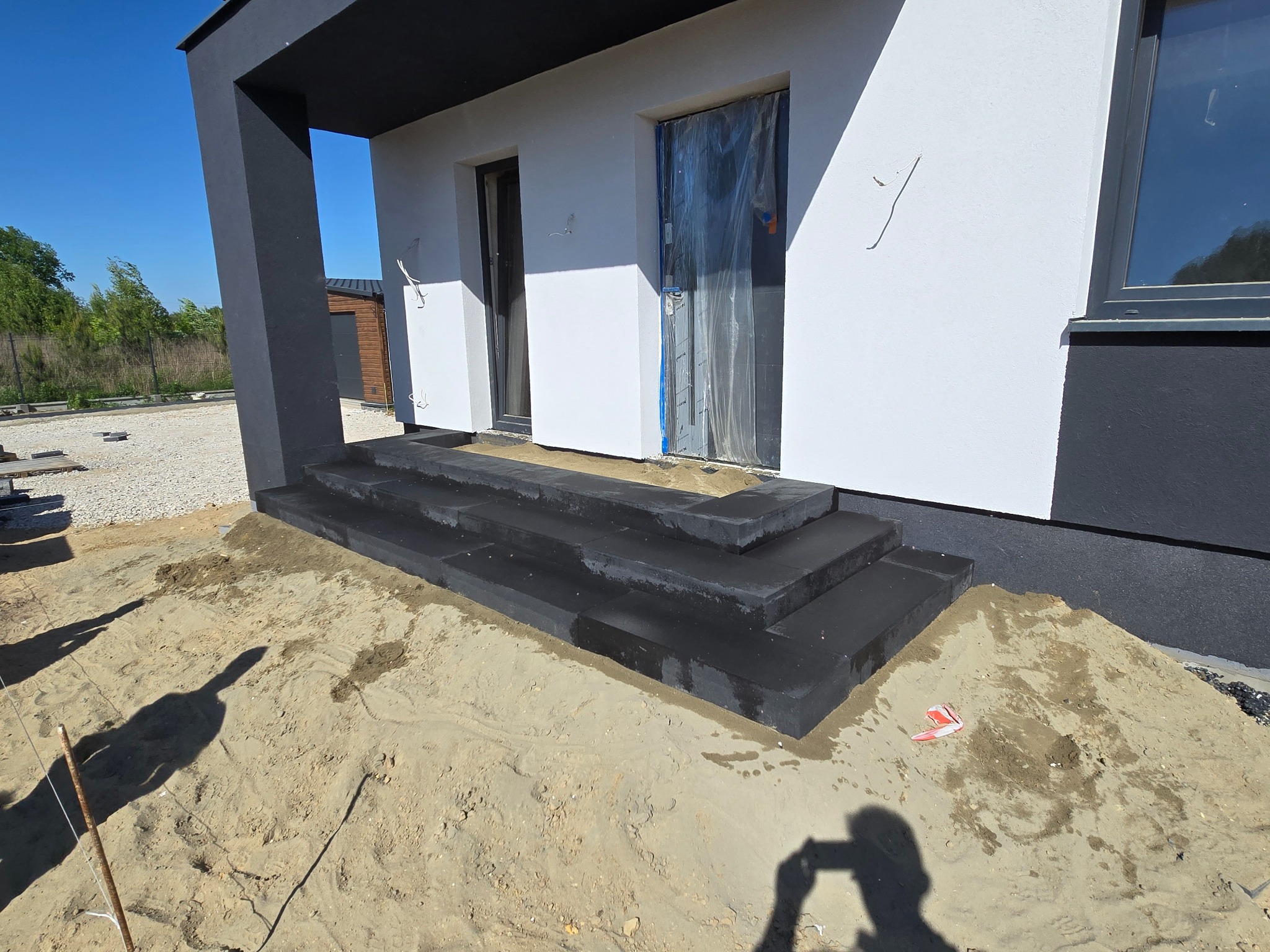 Nowoczesne schody kamienne z ciemnego granitu w Gorzkowicach, prowadzące do wejścia budynku. Widoczne świeżo ułożone stopnie i piasek. Budynek w bieli i szarości.