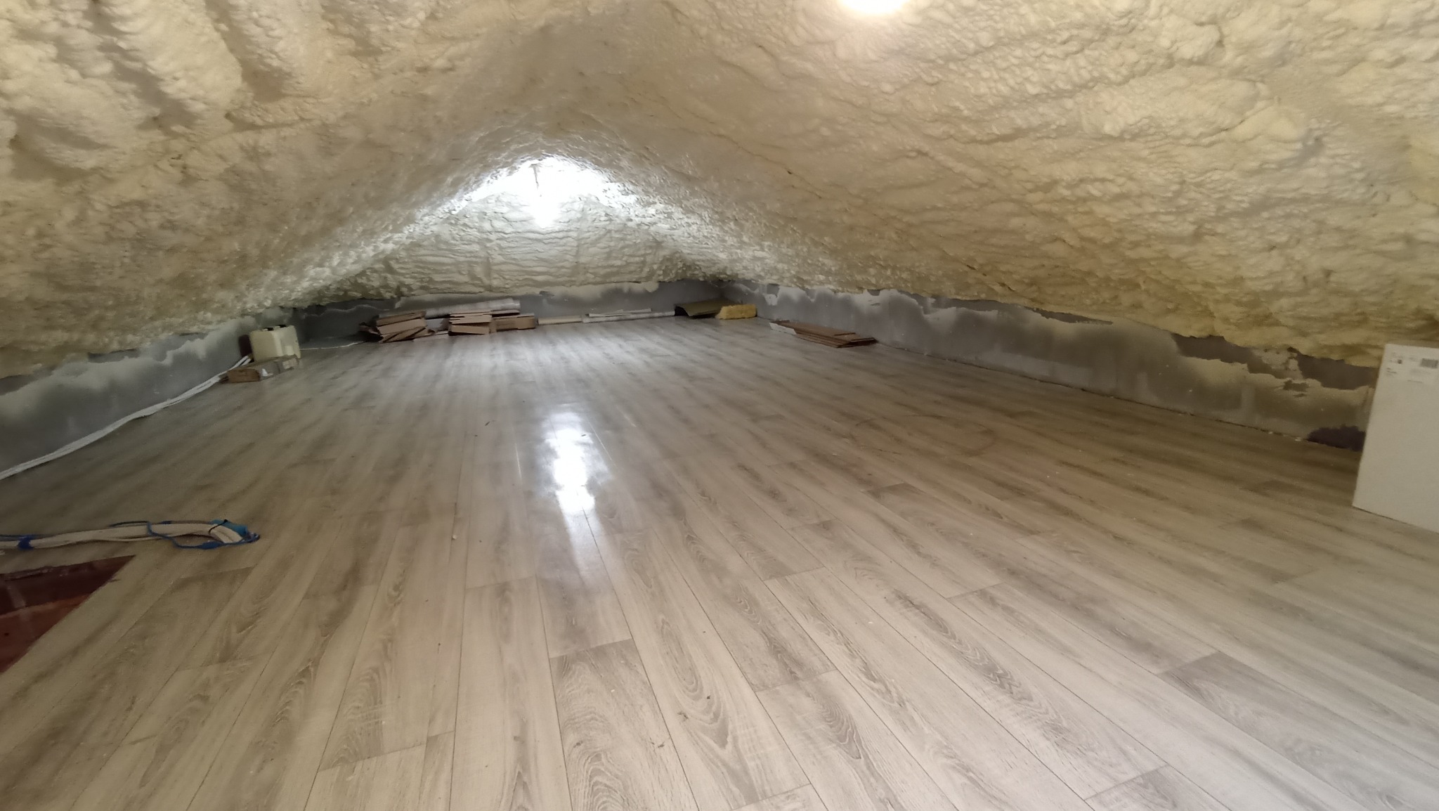 Poddasze po ociepleniu pianką: widoczne pokryte pianką ściany i sufit oraz jasna podłoga z paneli, z widocznym oknem dachowym w tle.