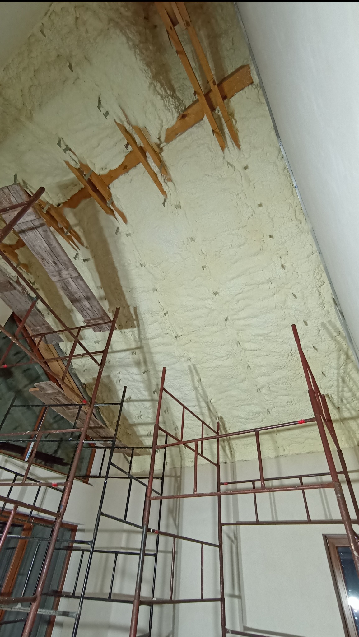 Niedokończone ocieplenie poddasza pianką poliuretanową, widoczne drewniane elementy konstrukcyjne i rusztowanie.