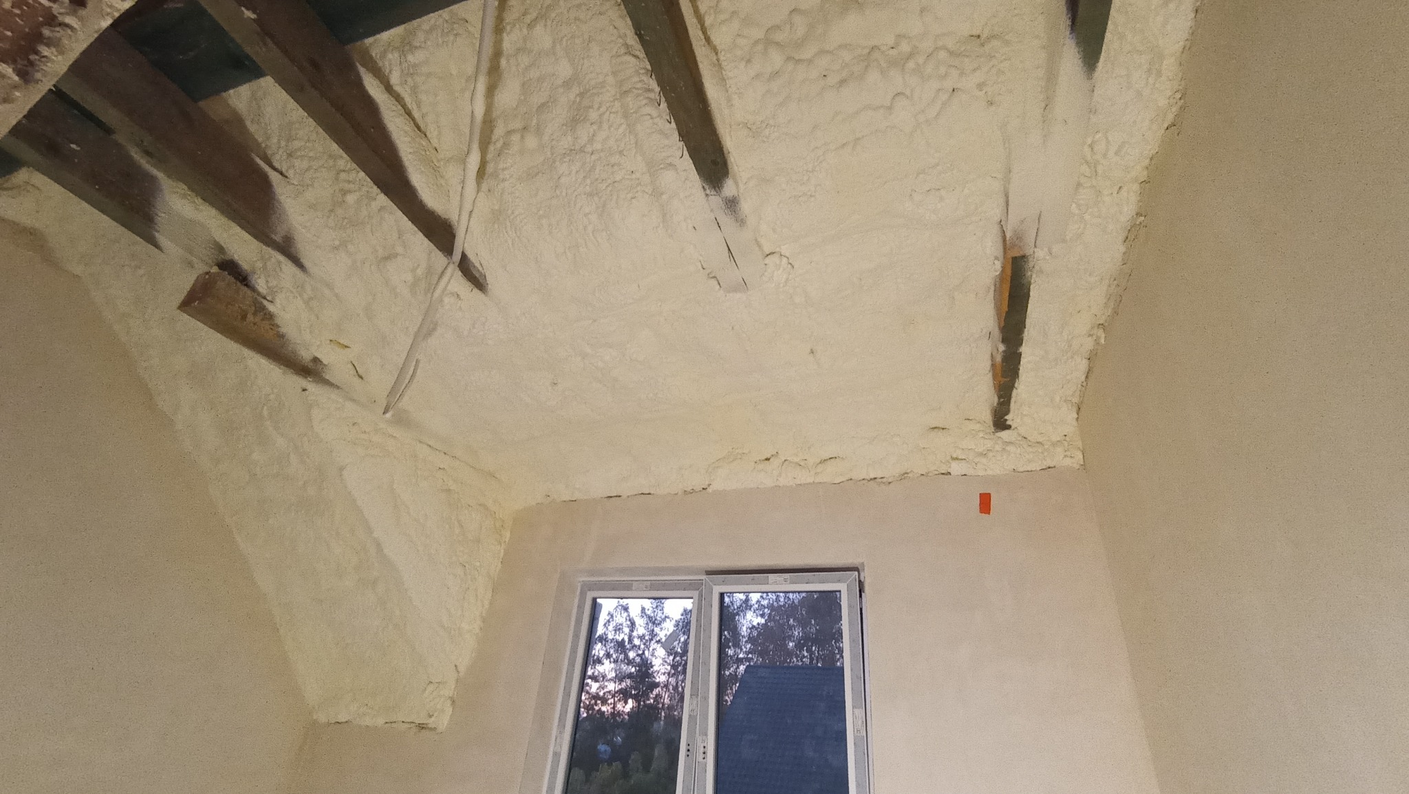 Poddasze z widoczną izolacją natryskową z pianki poliuretanowej (PUR), ukazującą nierównomierną strukturę i drewniane elementy konstrukcyjne dachu, widok na okno.