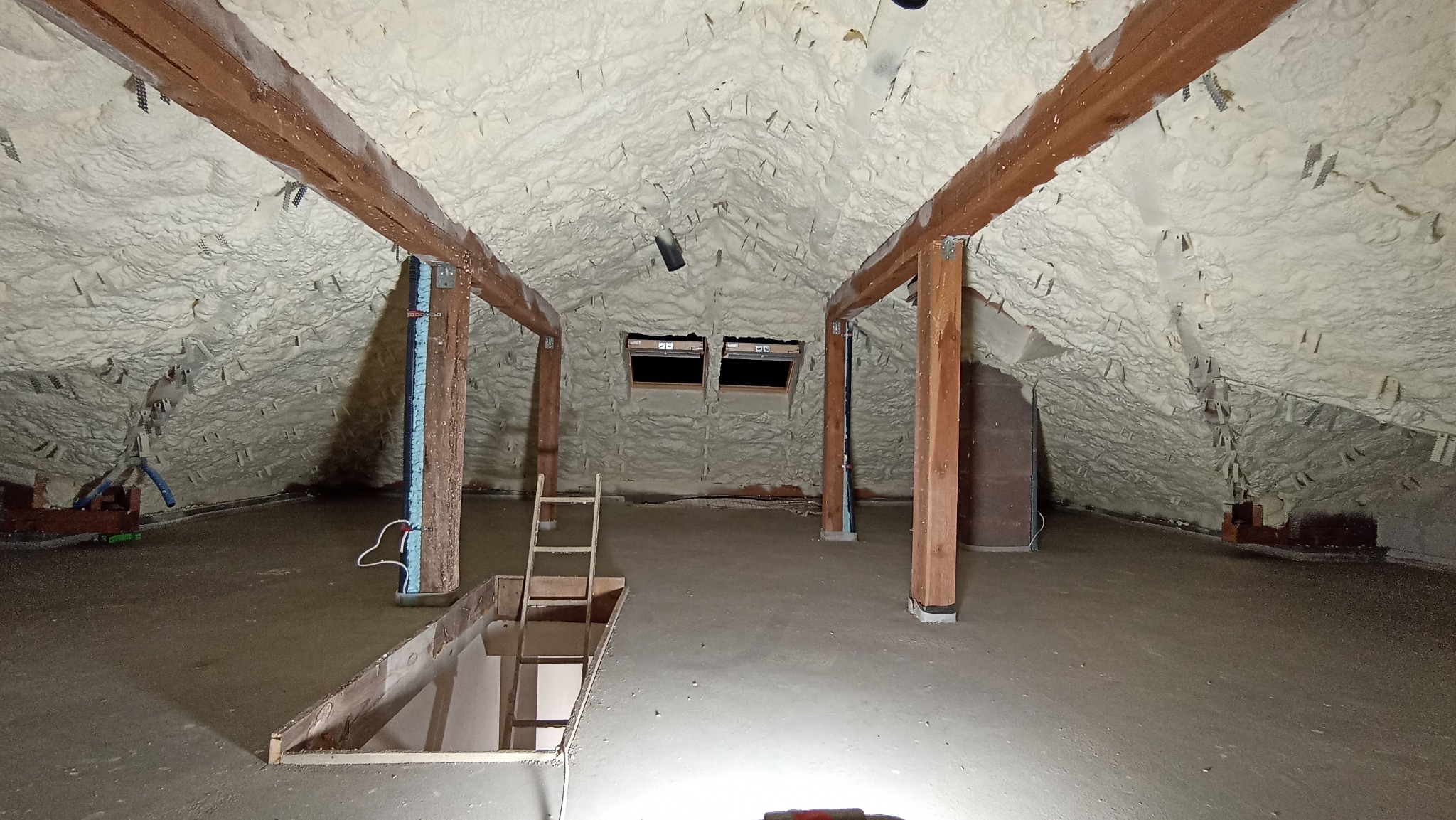 Poddasze po ociepleniu, pokryte pianką izolacyjną, z widocznymi drewnianymi belkami stropowymi i schodami prowadzącymi na dół. Na ścianach widoczne uchwyty montażowe. Dwa okna dachowe w tle.