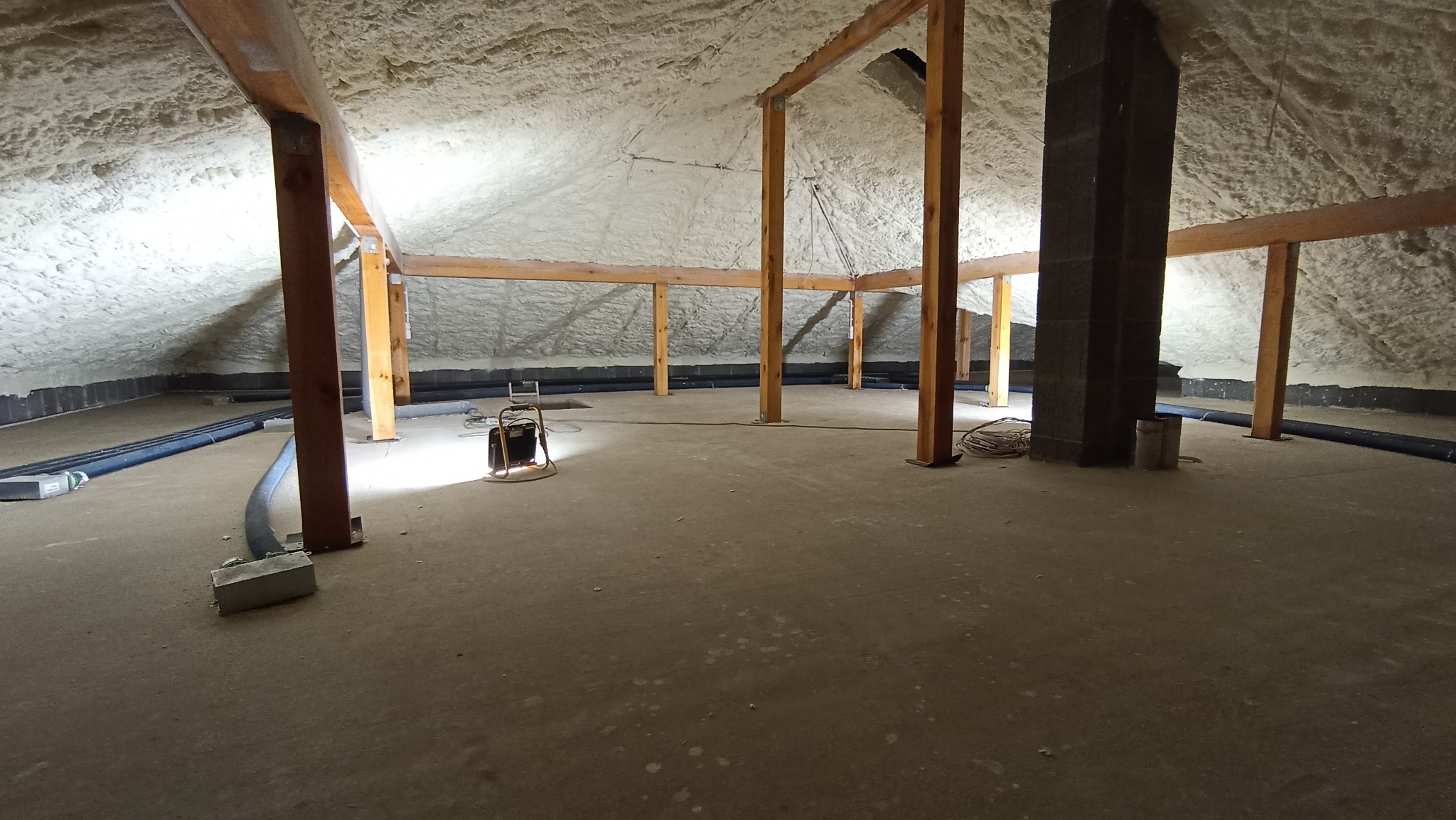 Poddasze po ociepleniu pianką: widoczne drewniane elementy konstrukcyjne, ciemny komin i szare węże wentylacyjne na podłodze, oświetlone lampą halogenową.