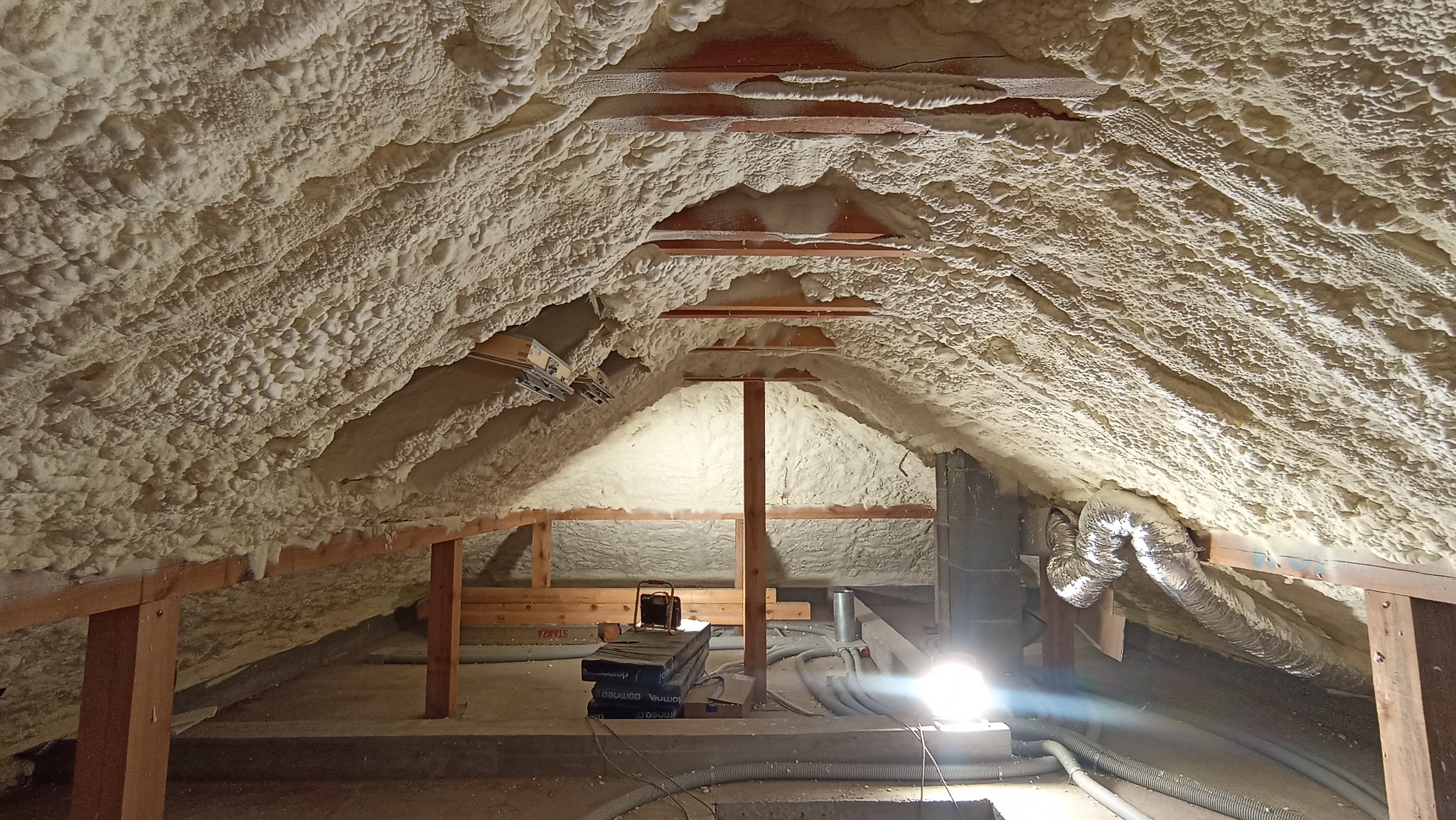 Poddasze ocieplone pianką, widoczne drewniane elementy konstrukcyjne, przewody wentylacyjne i lampa halogenowa na podłodze.