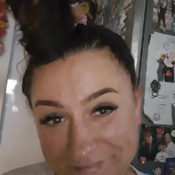 Portret kobiety z włosami upiętymi w kok, z widoczną lodówką z magnesami i zdjęciami w tle.