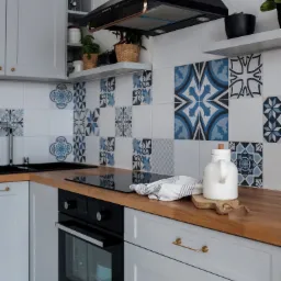 Kompaktowa kuchnia w odcieniach szarości z drewnianym blatem, ozdobnymi płytkami w stylu portugalskim i czarnym okapem, uzupełniona białym dzbankiem na desce do krojenia i roślinami w doniczkach...