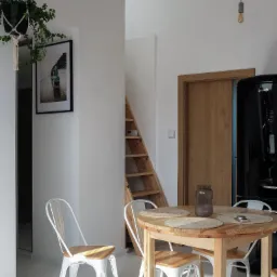 Jasne wnętrze jadalni z okrągłym drewnianym stołem nakrytym jutowymi podkładkami, białymi krzesłami w stylu industrialnym, czarną lodówką Gorenje i drewnianymi schodami w tle, doświetlone wiszącymi...