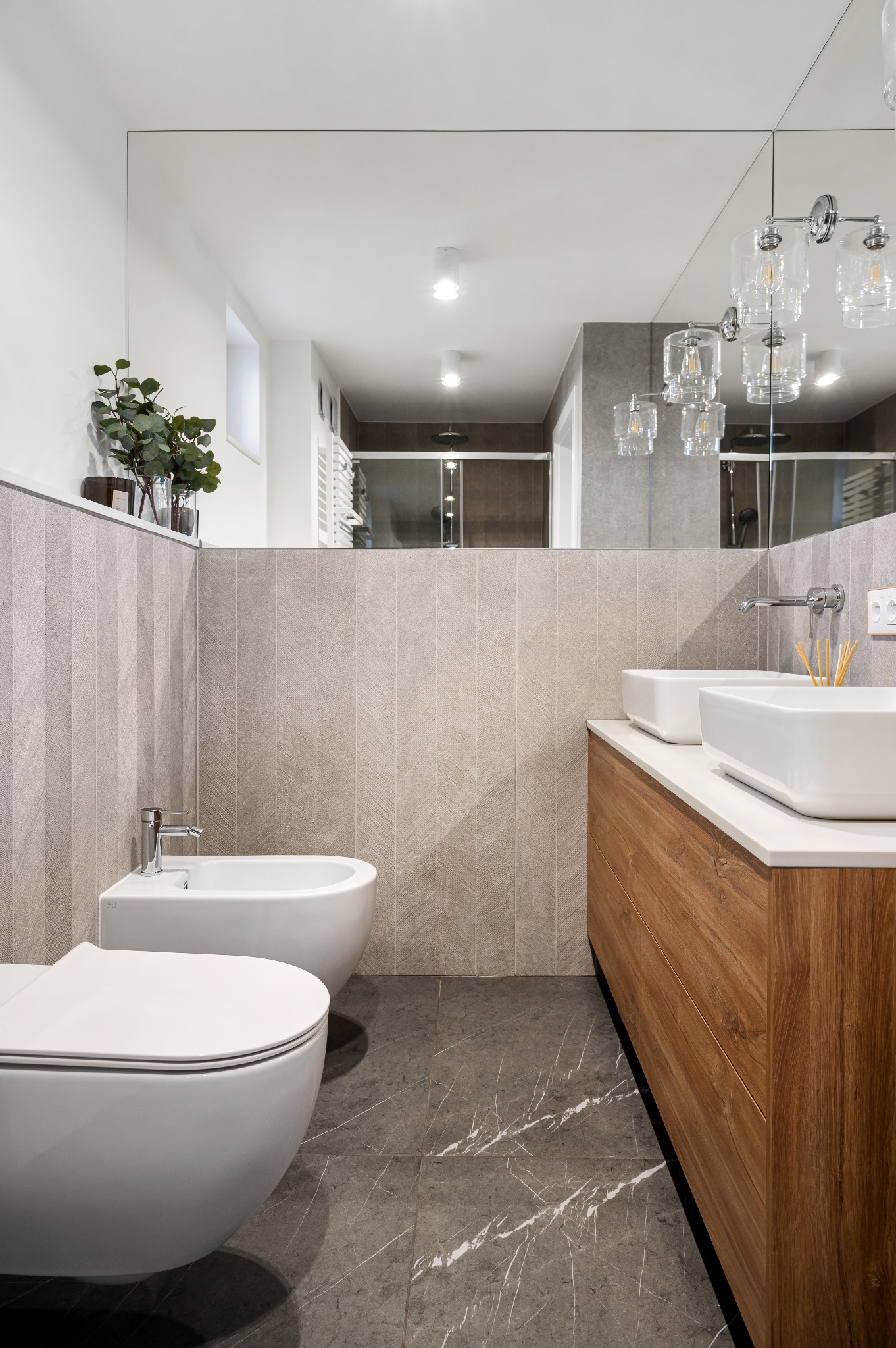 Nowoczesna łazienka z szarą marmurową podłogą, drewnianą szafką pod umywalki, białymi umywalkami nablatowymi, wiszącą toaletą i bidetem. Na ścianie płytki w pionowe, subtelne wzory. Duże lustro...