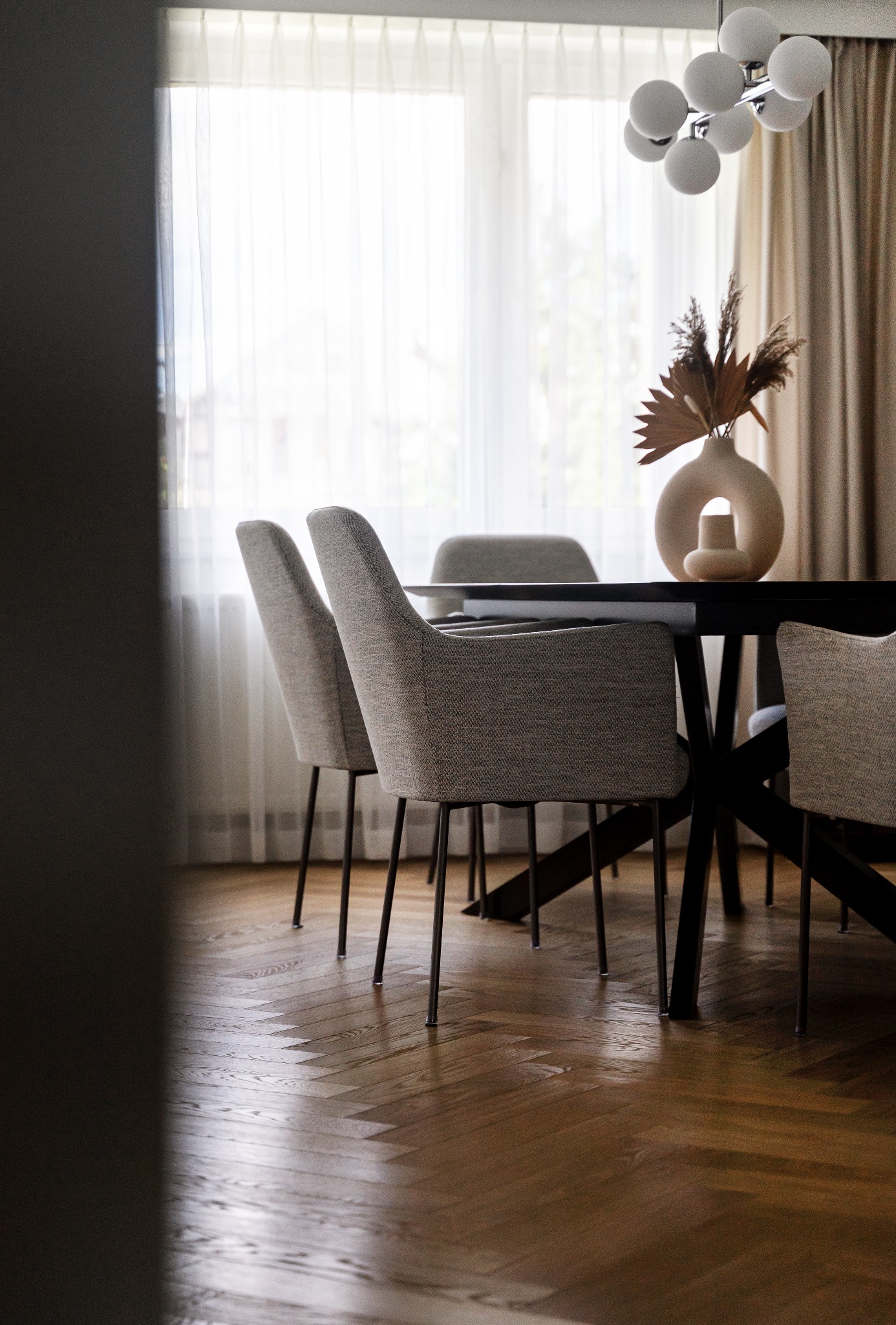 Jadalnia z ciemnym stołem, krzesłami w odcieniach szarości, parkietem w jodełkę i nowoczesną lampą sufitową z kulistymi kloszami, w tle okno z białymi zasłonami.