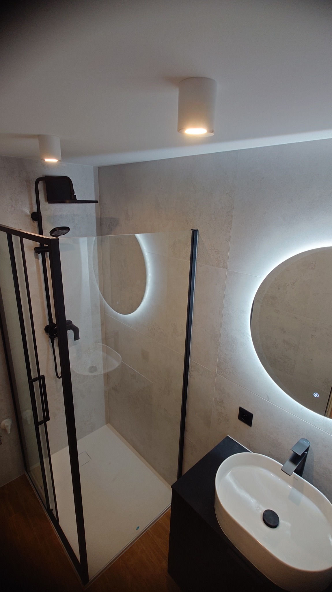Nowoczesna łazienka z kabiną prysznicową typu walk-in z czarnymi ramami, okrągłym lustrem z podświetleniem LED i umywalką nablatową. Minimalistyczny design, szare płytki.