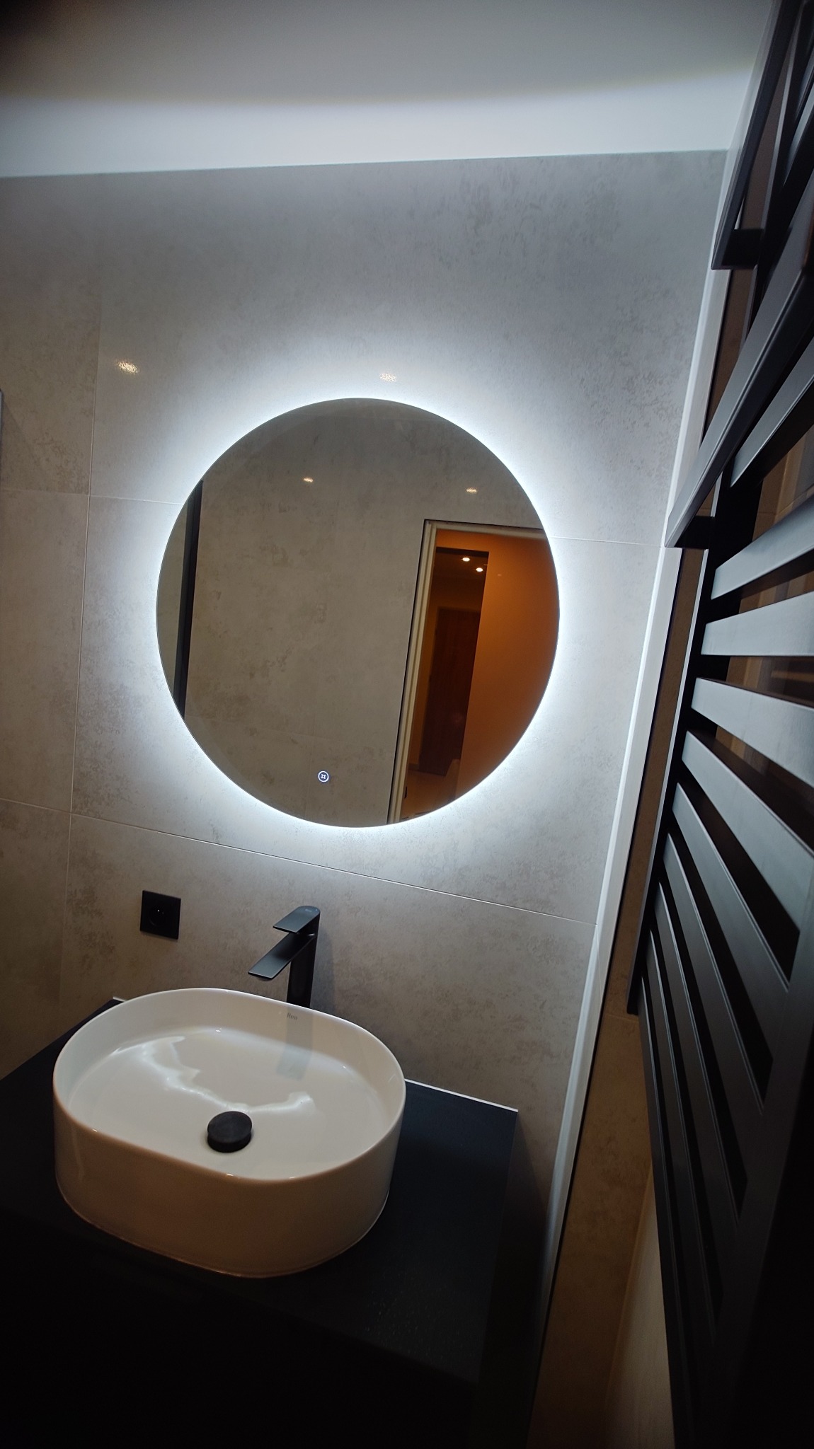 Nowoczesna łazienka z okrągłym lustrem LED, białą umywalką nablatową i czarną armaturą. Szare płytki na ścianach i grzejnik drabinkowy w ciemnym kolorze.