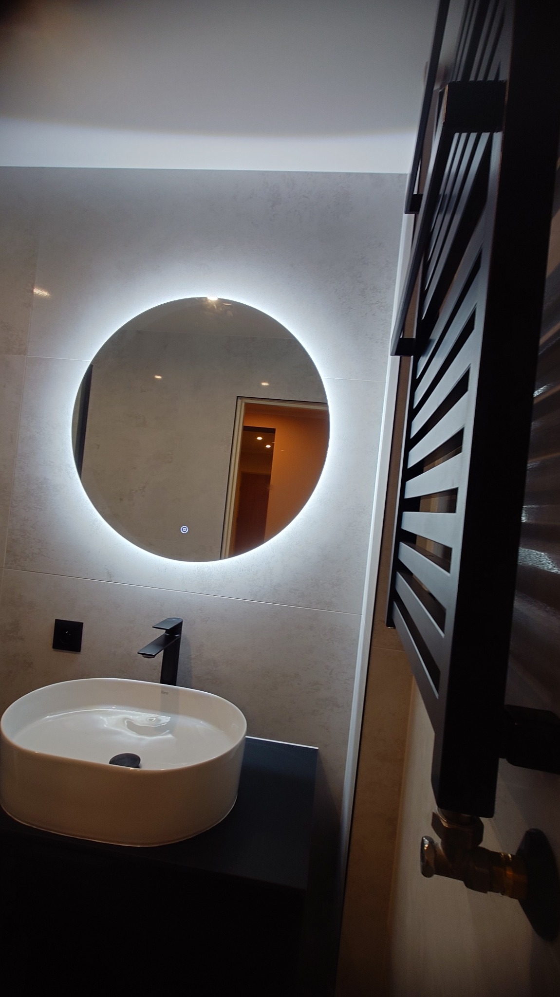 Nowoczesna łazienka: owalne lustro z podświetleniem LED, biała umywalka nablatowa, czarna bateria i grzejnik drabinkowy. Minimalistyczny design, szare płytki.