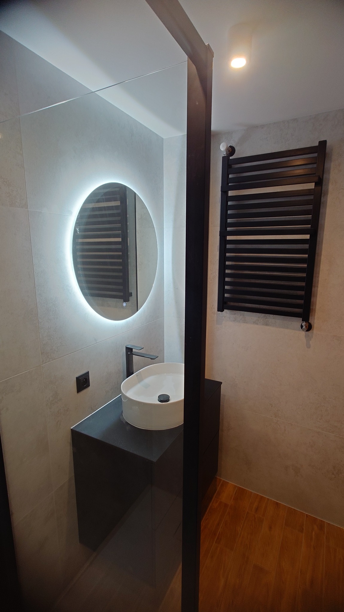 Nowoczesna łazienka z okrągłym lustrem LED, białą umywalką na czarnej szafce i czarnym grzejnikiem drabinkowym. Jasne płytki i drewnopodobna podłoga.