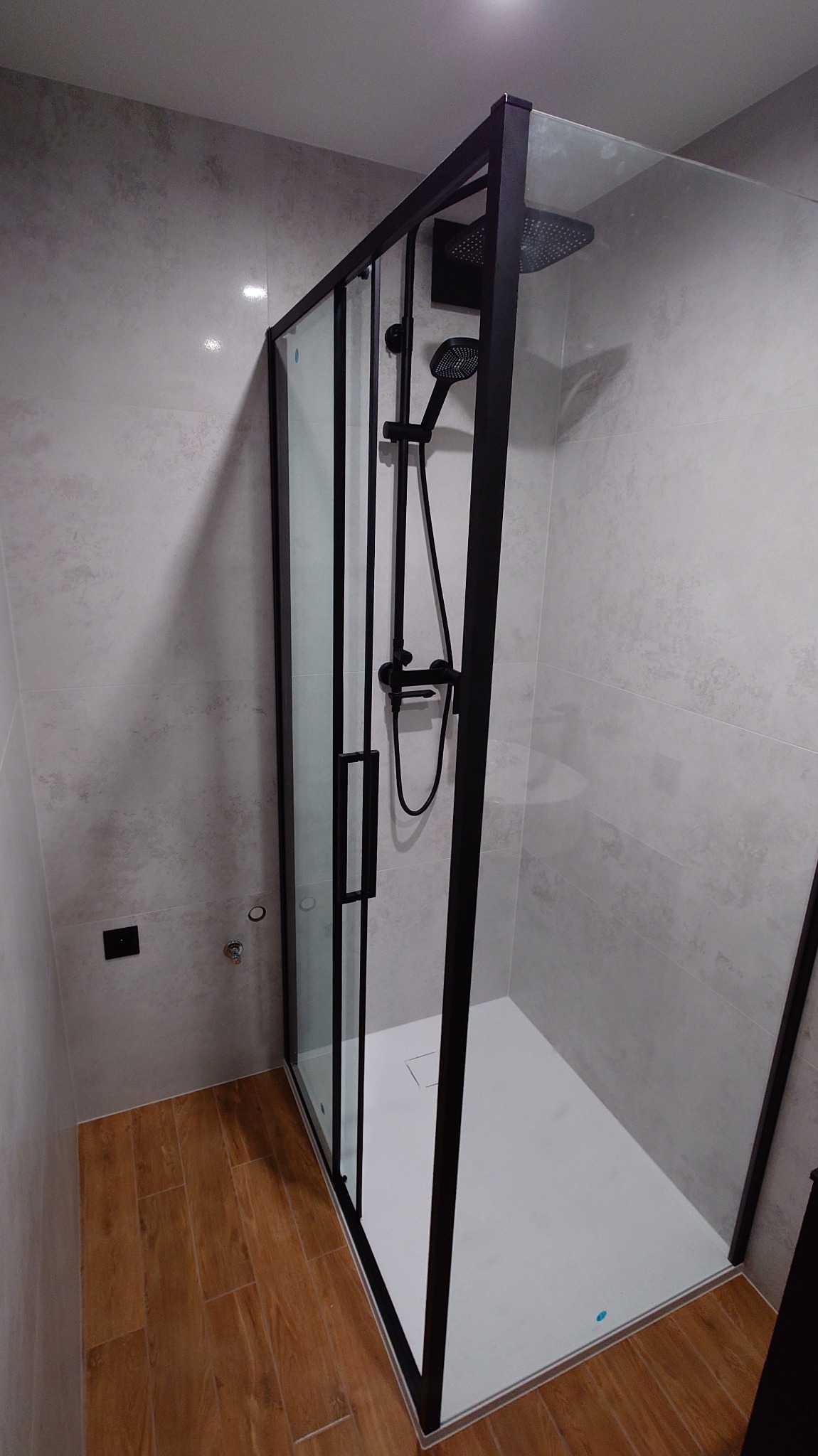 Nowoczesna kabina prysznicowa z czarnymi profilami i deszczownicą, na tle szarych płytek i drewnopodobnej podłogi. Minimalistyczny design, widok z boku.