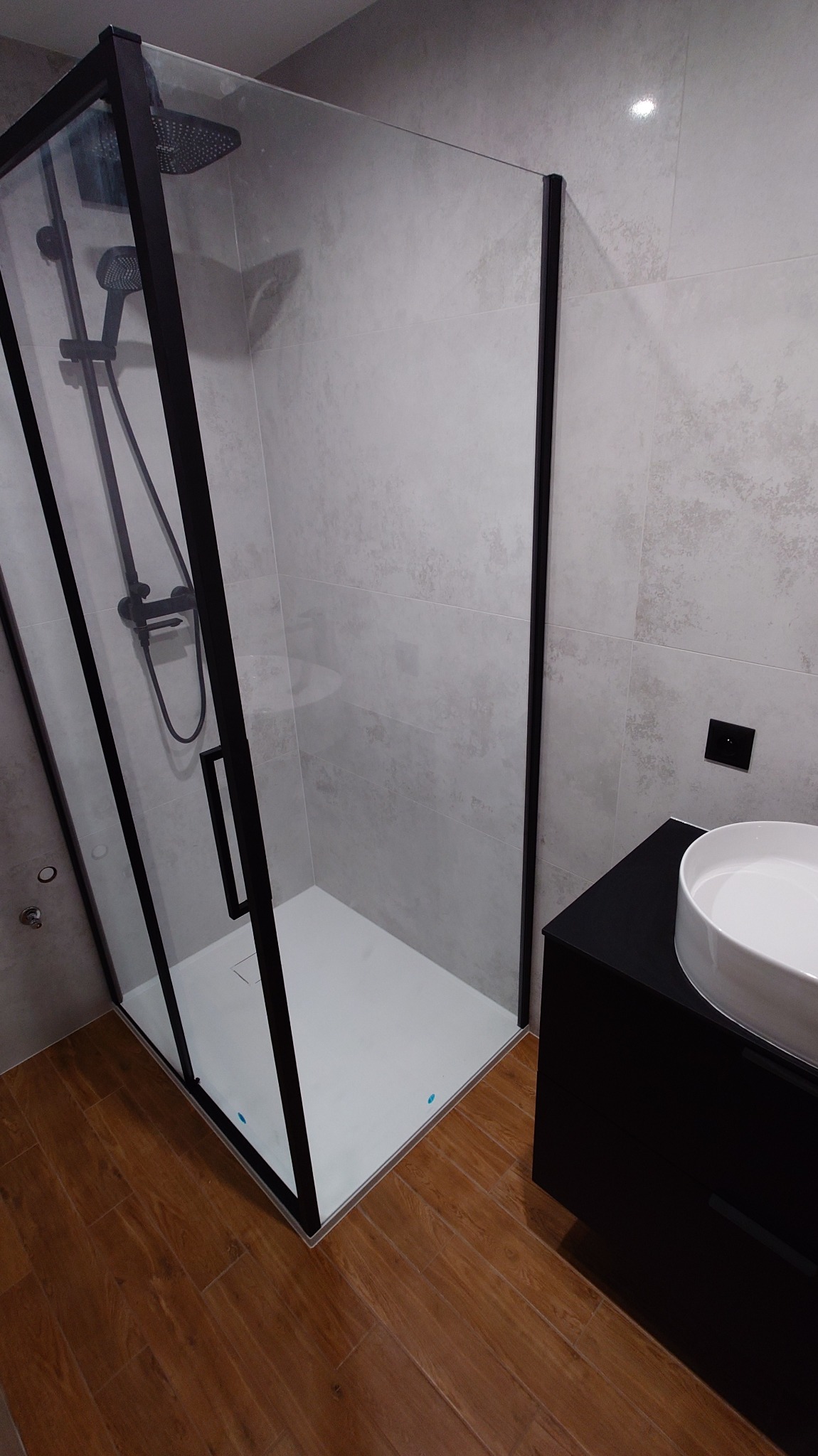 Nowoczesna kabina prysznicowa z czarną ramą i deszczownicą, jasne płytki na ścianach, drewnopodobna podłoga, umywalka nablatowa na czarnej szafce.