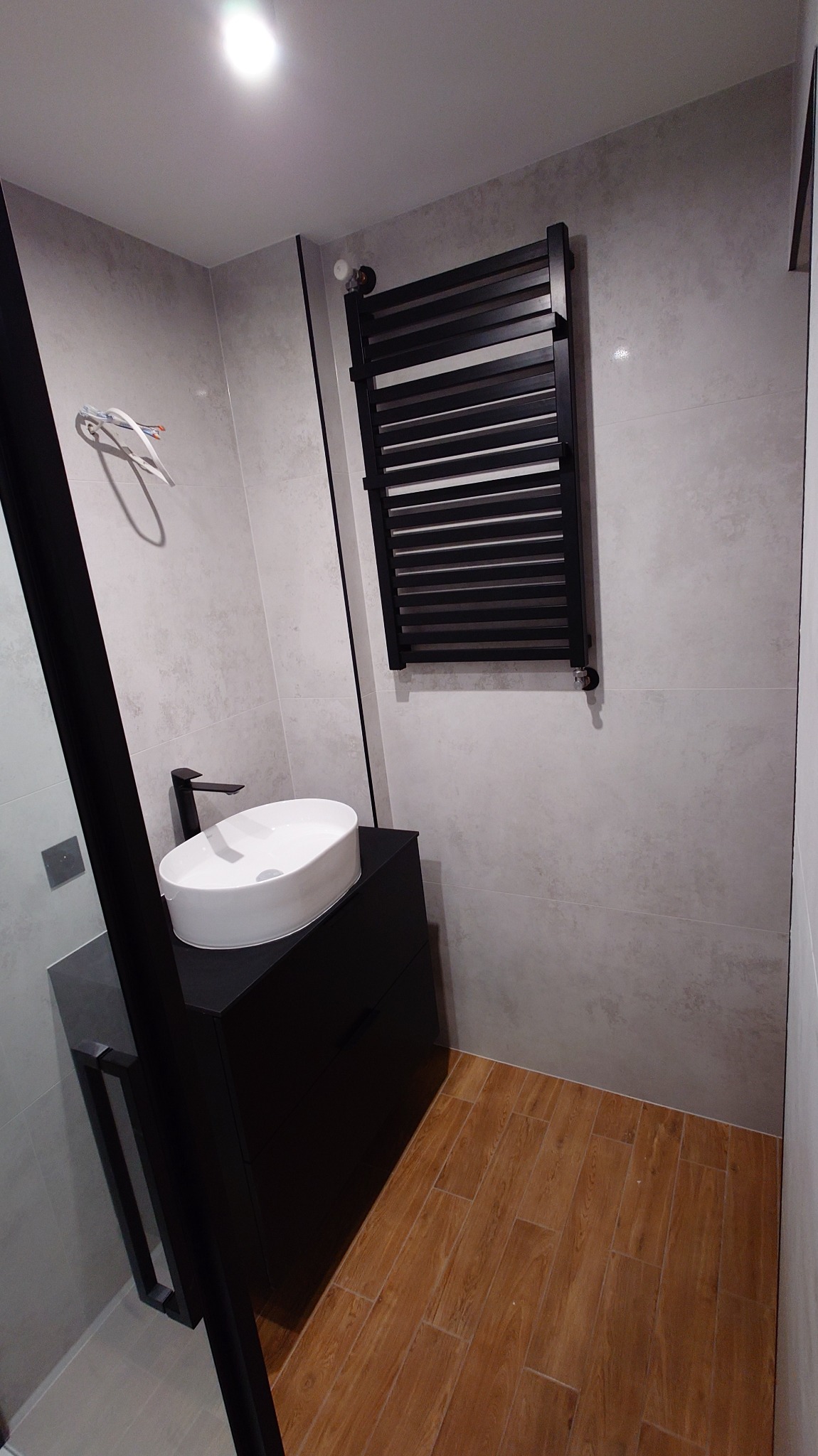 Nowoczesna łazienka z drewnopodobną podłogą, umywalką nablatową na czarnej szafce i czarnym grzejnikiem drabinkowym na tle szarych płytek.