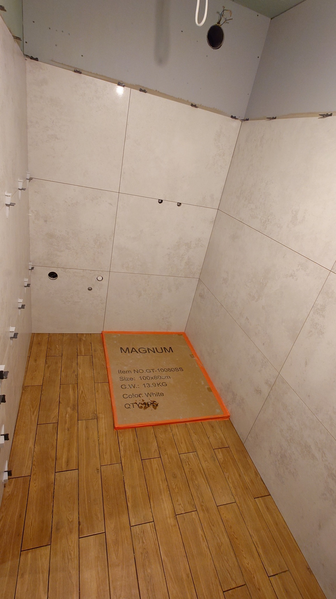 Łazienka w trakcie wykańczania: beżowe płytki na ścianach z krzyżykami dystansowymi, drewnopodobne panele na podłodze, karton z informacjami o płytkach.