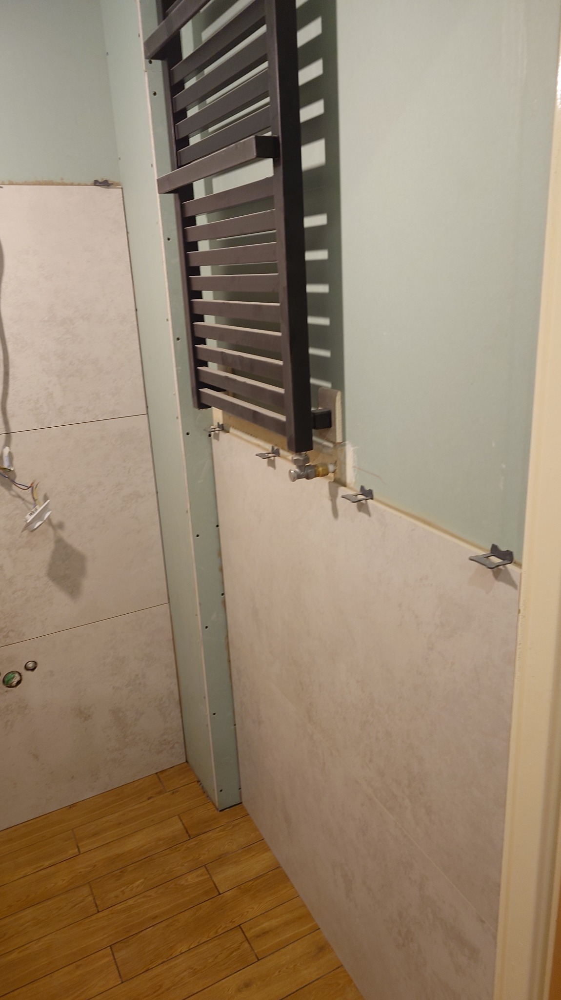 Montaż płytek w łazience z grzejnikiem drabinkowym. Widoczne płytki na ścianie, drewnopodobna podłoga i elementy instalacji grzewczej. Etap prac wykończeniowych.