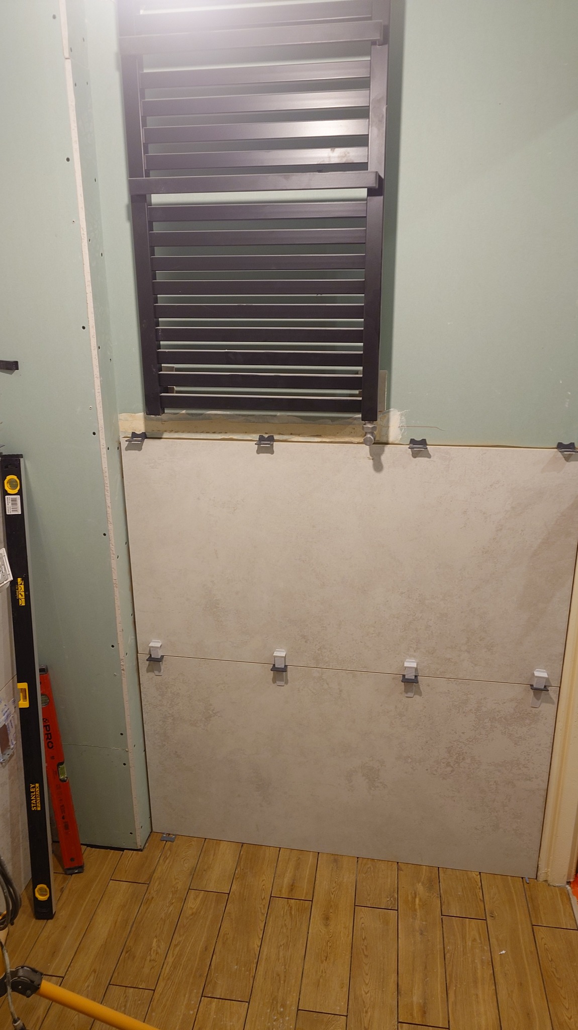 Montaż płytek ściennych w łazience, widoczne klipsy poziomujące, grzejnik drabinkowy i poziomica. Podłoga wyłożona panelami imitującymi drewno.