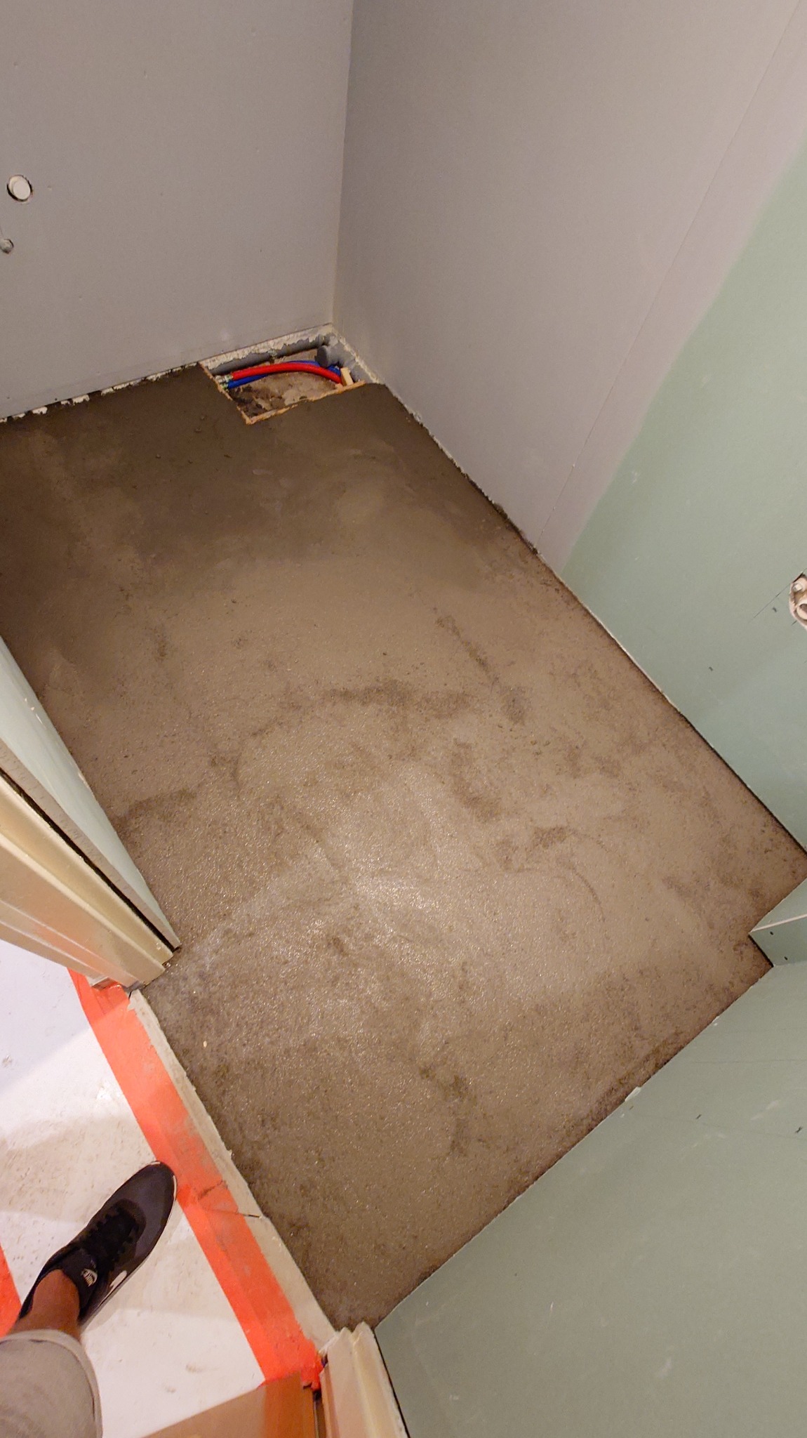 Wylany, świeży podkład podłogowy w rogu łazienki, z widocznymi rurami instalacji w narożniku. Ściany przygotowane do dalszych prac wykończeniowych.