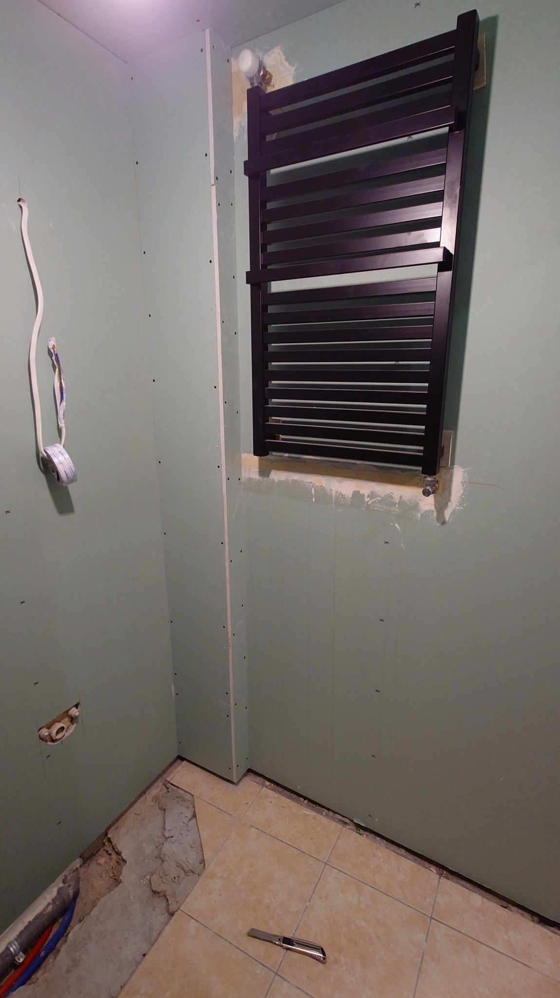 Montaż czarnego grzejnika drabinkowego na ścianie z płyt gipsowo-kartonowych w trakcie wykańczania łazienki. Widoczne narzędzie i fragment podłogi z uszkodzonym fragmentem.