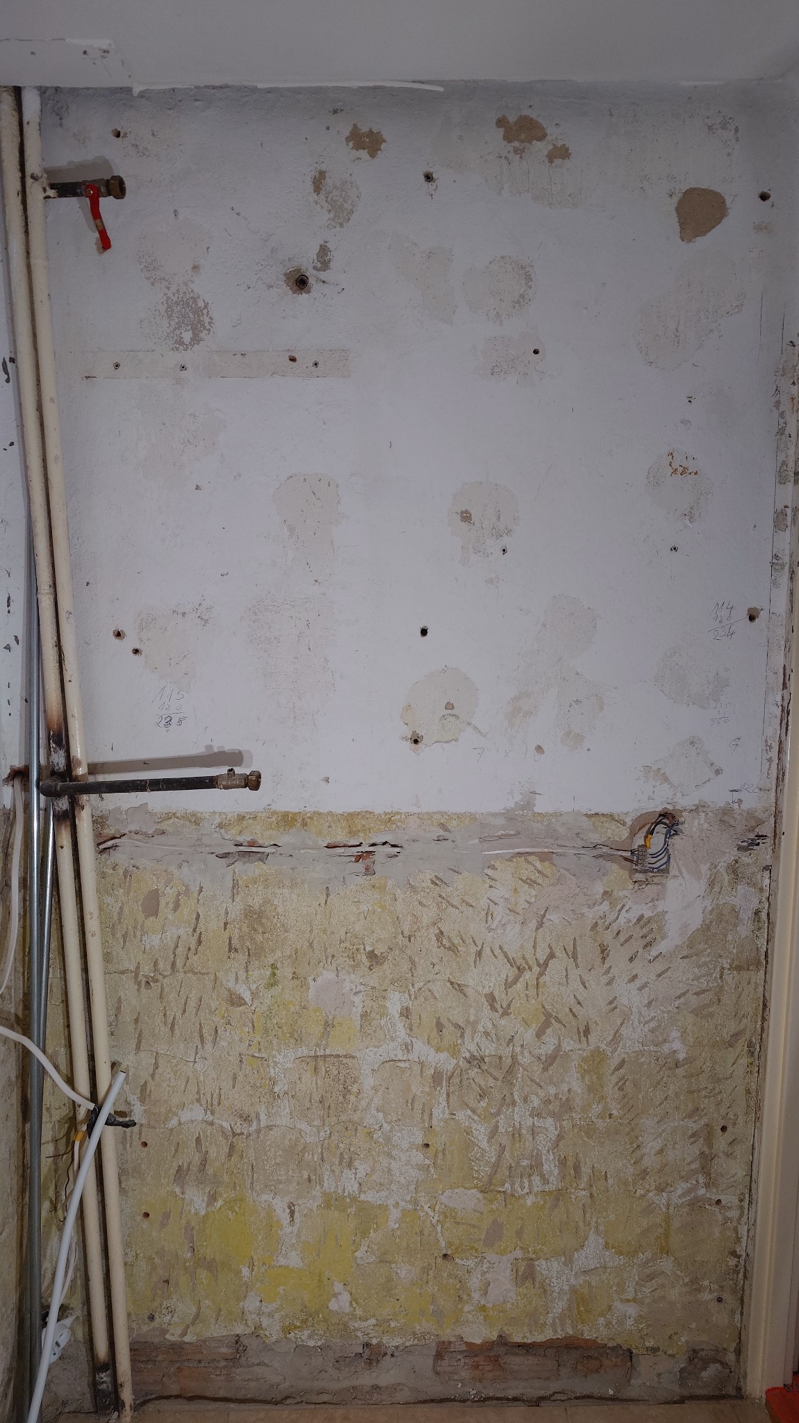 Ściana w trakcie przygotowania do remontu: widoczne rury, instalacja elektryczna i ślady po starych płytkach. Częściowo usunięty tynk odsłania strukturę ściany.