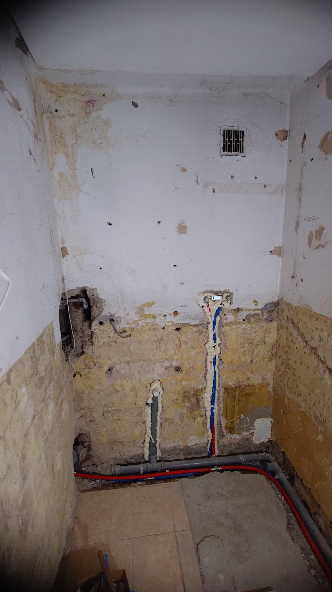 Wnętrze łazienki w trakcie remontu: odsłonięte ściany z instalacjami wodnymi (czerwone i niebieskie rury) zabezpieczonymi pianką montażową, widoczne ślady po płytkach.