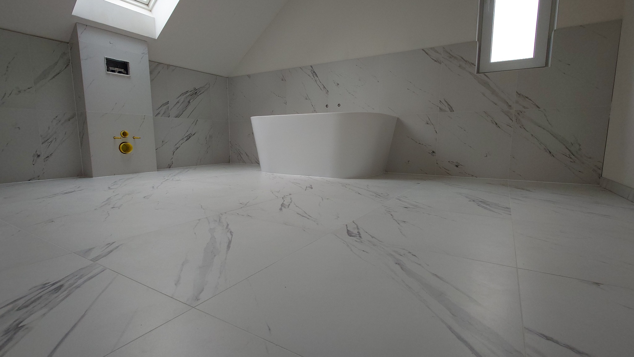 Nowoczesna łazienka na poddaszu z białą wanną wolnostojącą i marmurowymi płytkami na podłodze i ścianach. Okno dachowe wpuszcza naturalne światło.