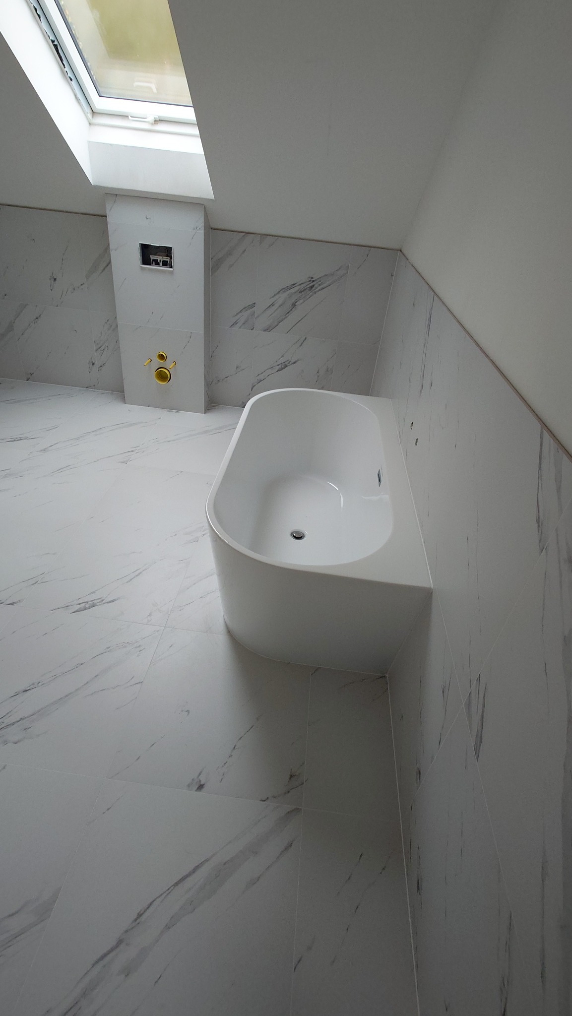 Biała wanna w nowoczesnej łazience z marmurowymi płytkami i oknem dachowym. Minimalistyczny design, jasne wnętrze, widoczne elementy instalacji.