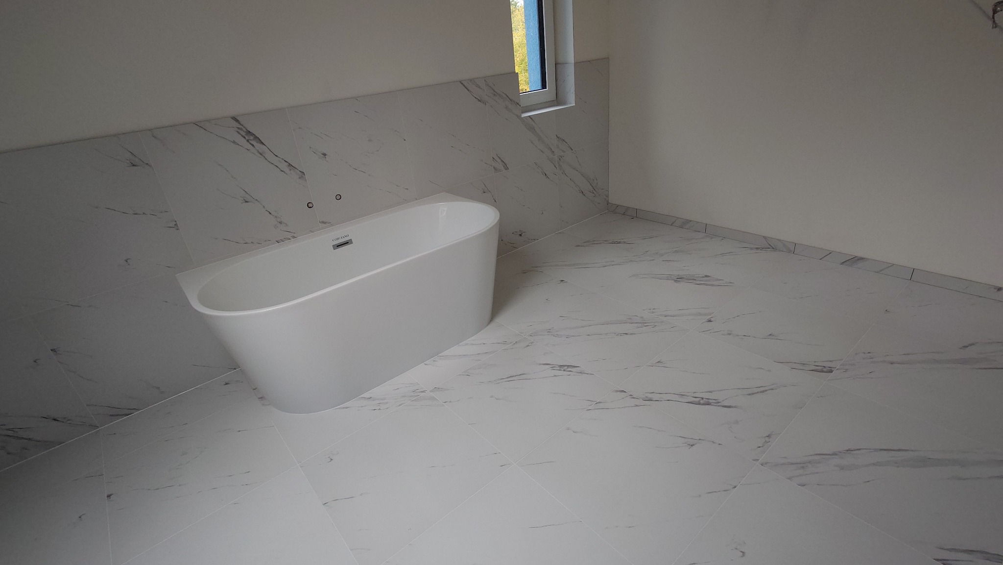 Nowoczesna łazienka z wolnostojącą białą wanną i marmurowymi płytkami na podłodze i ścianach. Minimalistyczny design, okno w tle.