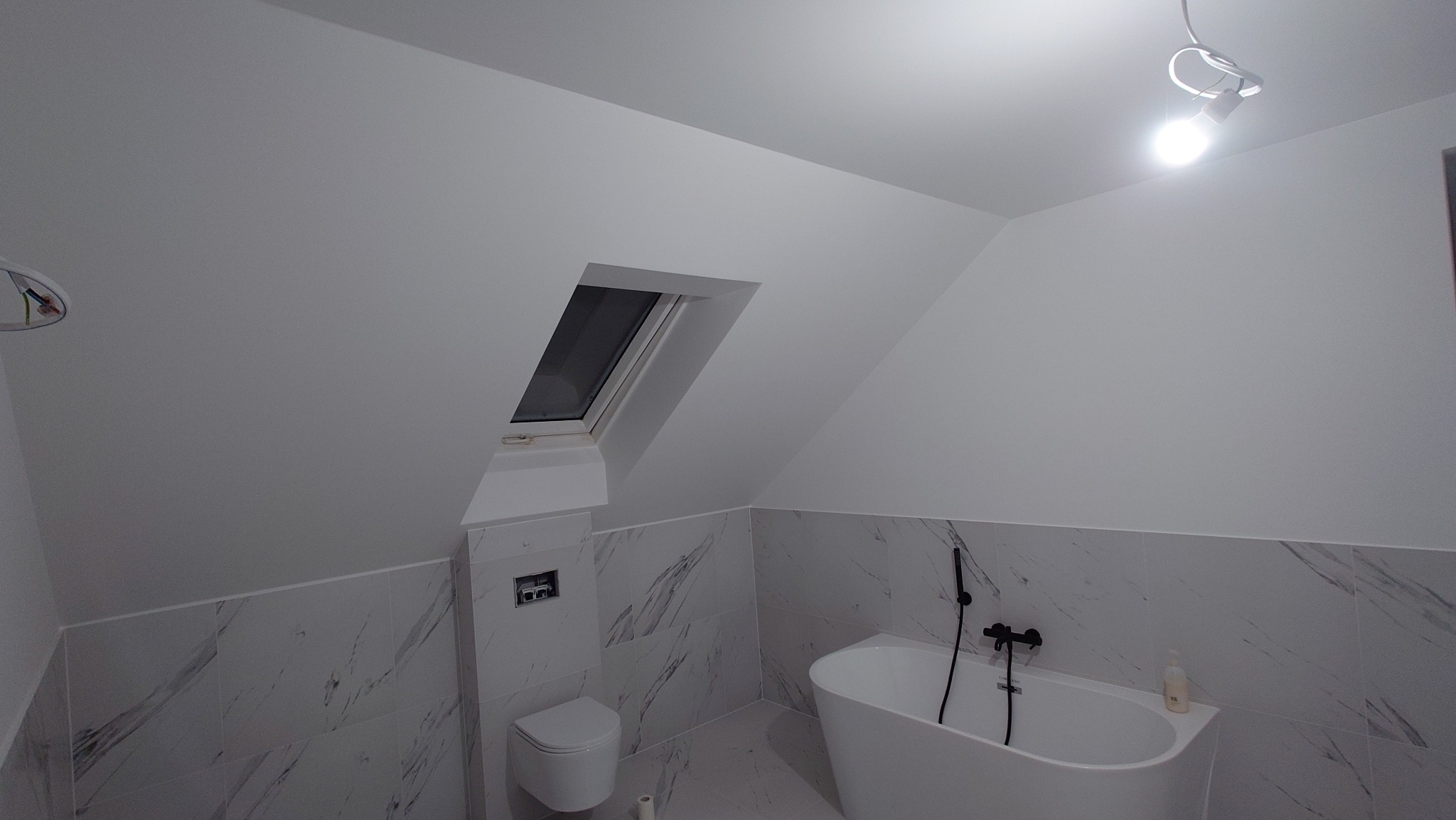 Wykończona łazienka na poddaszu z oknem dachowym, białą wanną wolnostojącą i marmurowymi płytkami. Minimalistyczny styl, czarne elementy armatury.